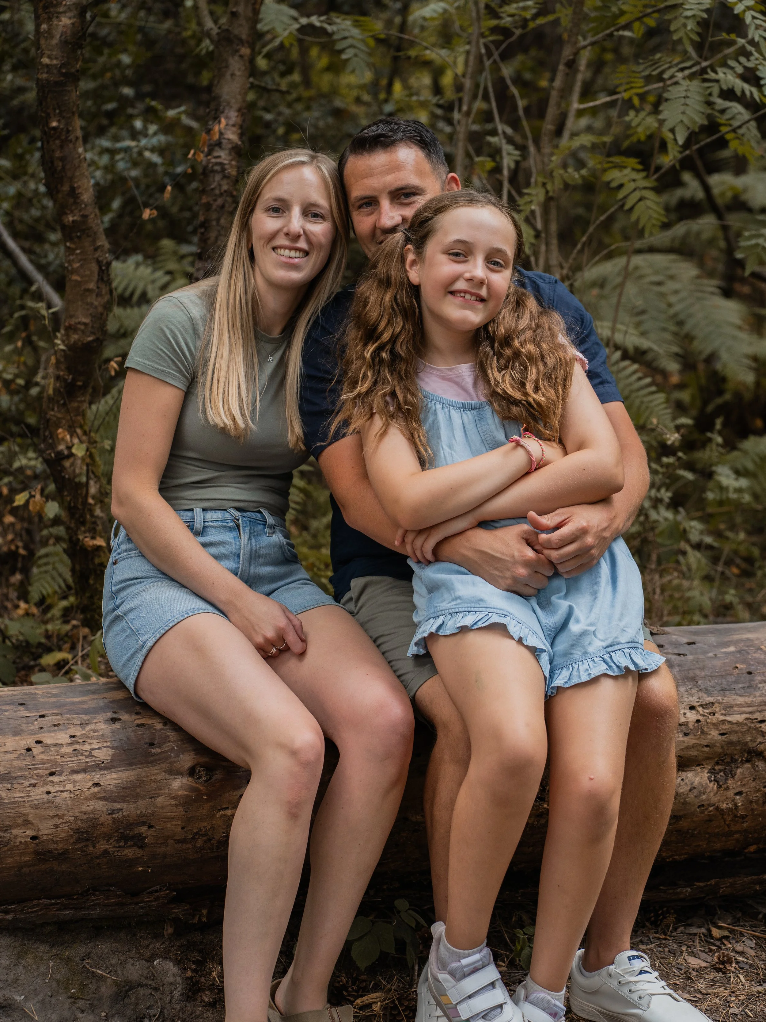 A family of three sitting closely together on a fallen log in a wooded area, smiling at the camera. The mother has long blonde hair, the father has short dark hair, and the daughter has long, curly red hair and is sitting in front of her parents with