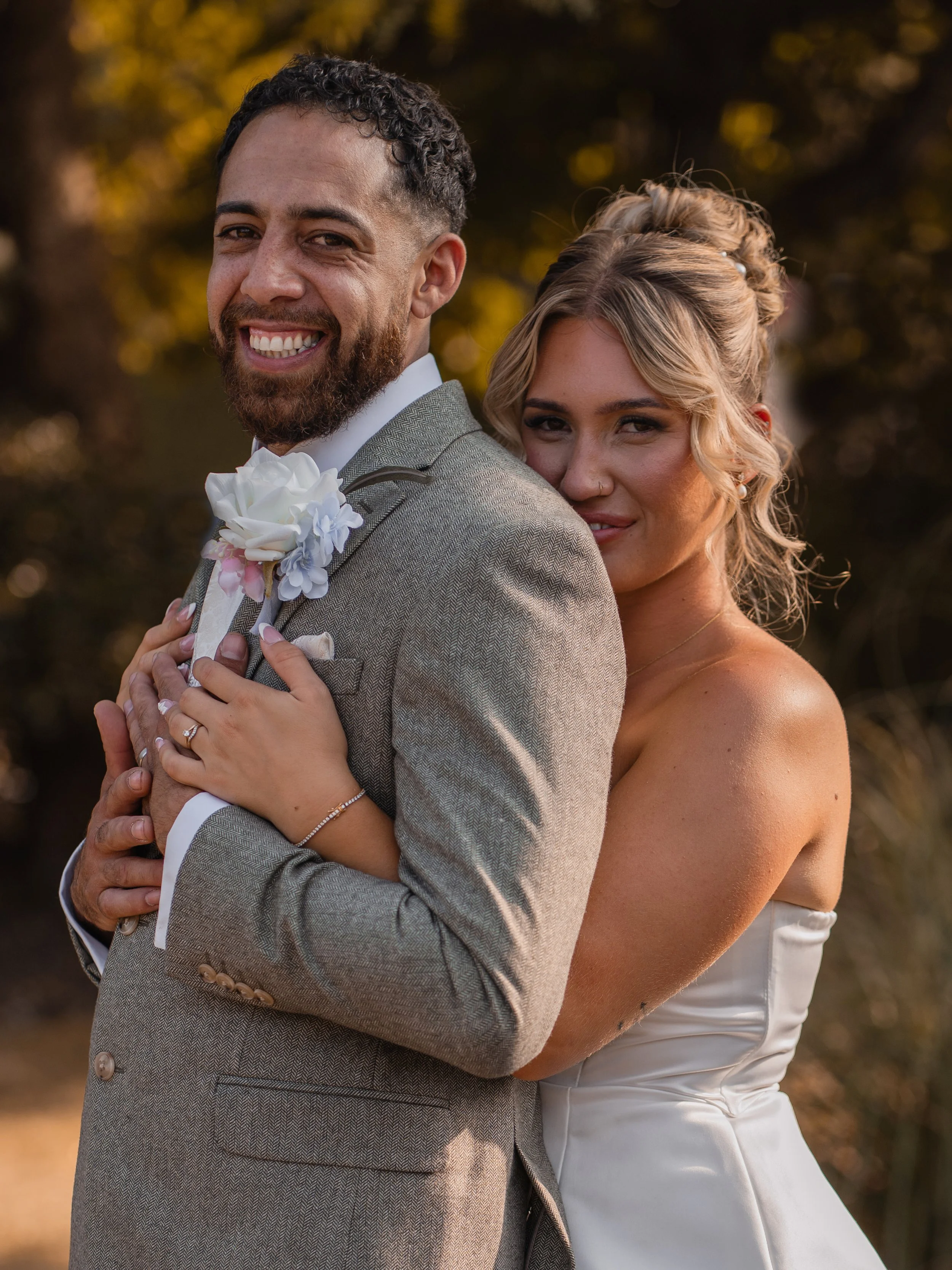 A smiling man in a suit and a woman in a white dress are embracing outdoors, with trees in the background.