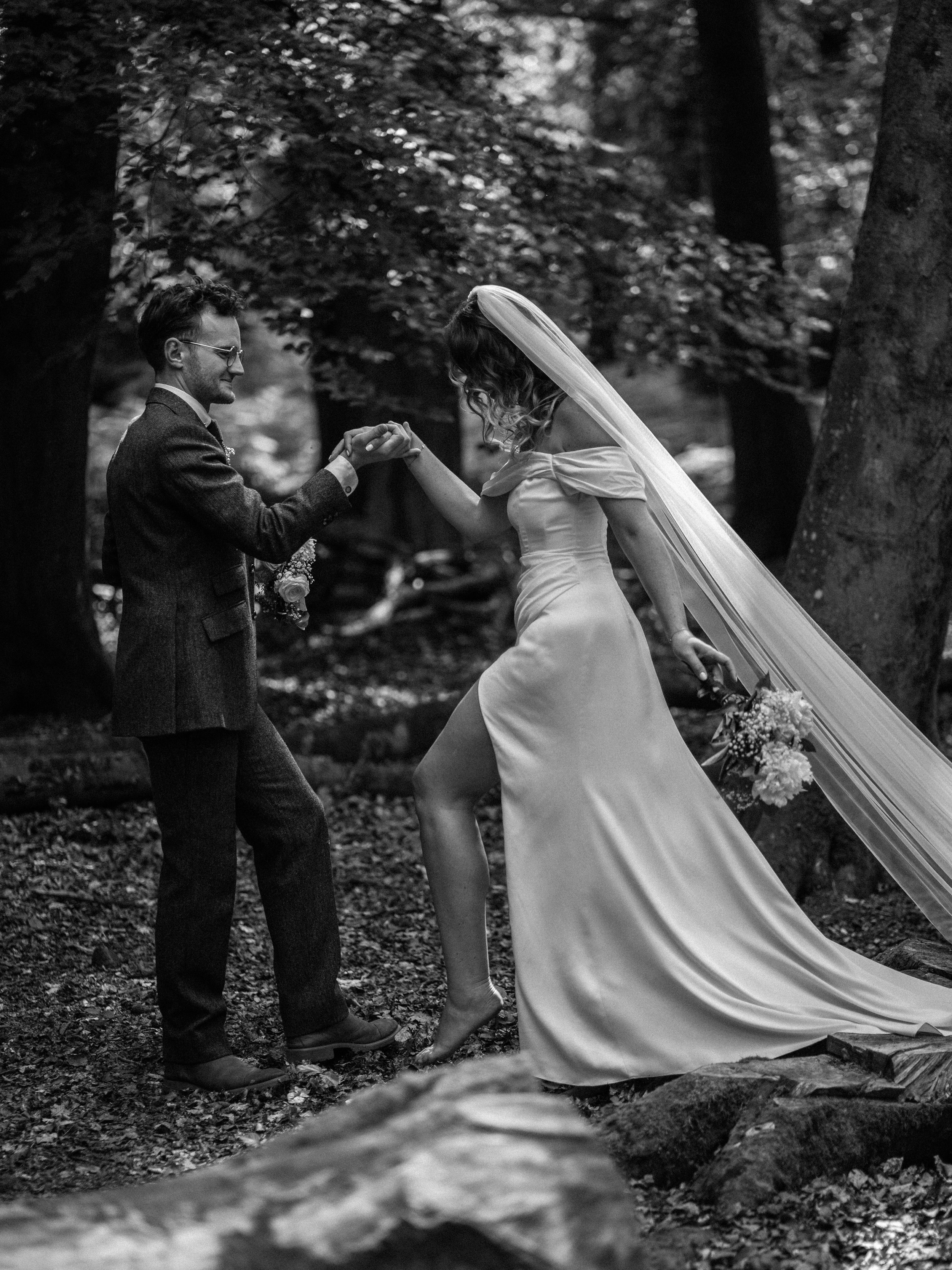 A black-and-white photo of a bride and groom holding hands in a forest. The woman is wearing a wedding dress and veil, and the man is in a suit. They are interacting lovingly, surrounded by trees and fallen leaves.