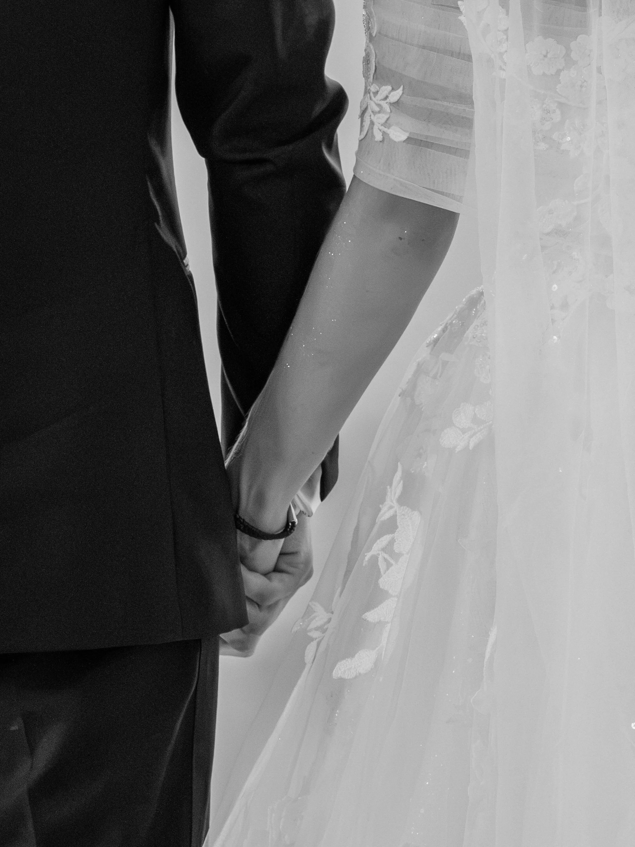 Close-up of two people holding hands. One person wears a black suit, and the other wears a wedding dress with lace details.