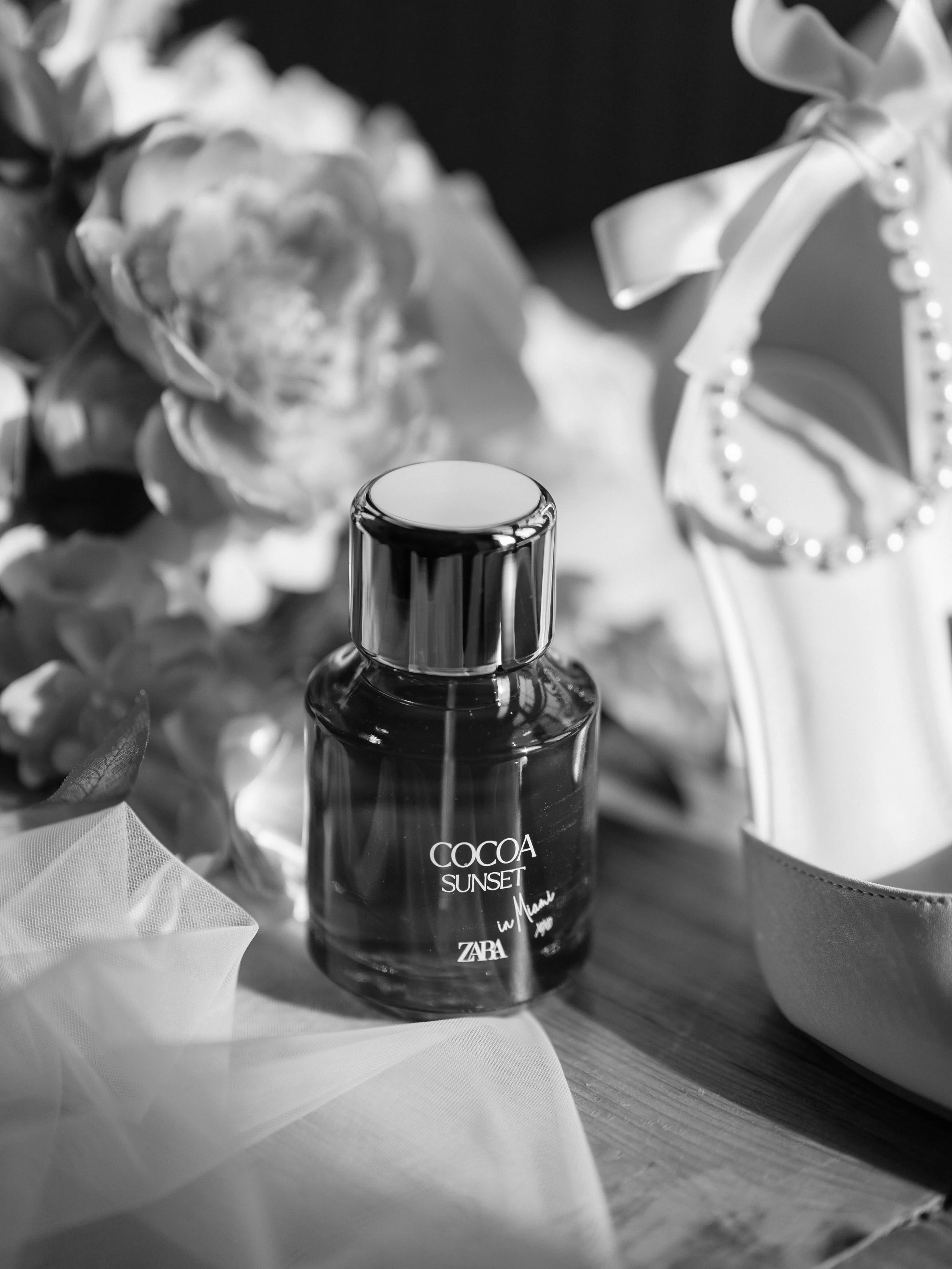 A perfume bottle labeled 'Cocoa Sunset' by Zara surrounded by flowers and fabric on a wooden surface.