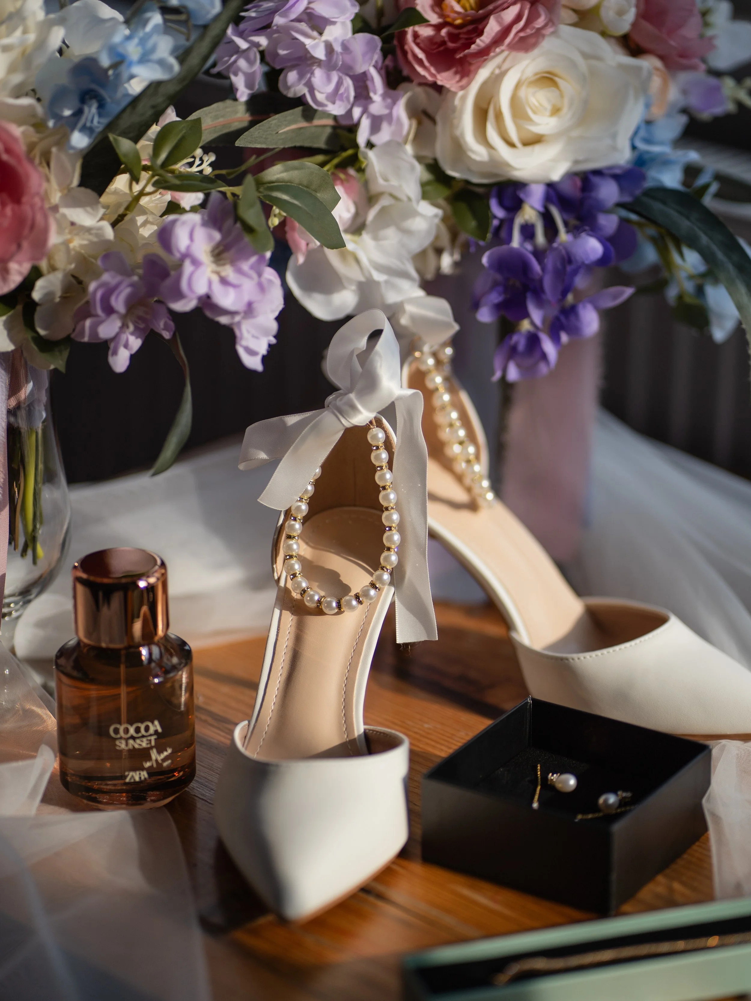 A close-up of a vanity table with a floral arrangement, pearl jewelry, a pair of white high heels, a brown perfume bottle labeled 'COCOA SUNSET,' and jewelry in a black box.