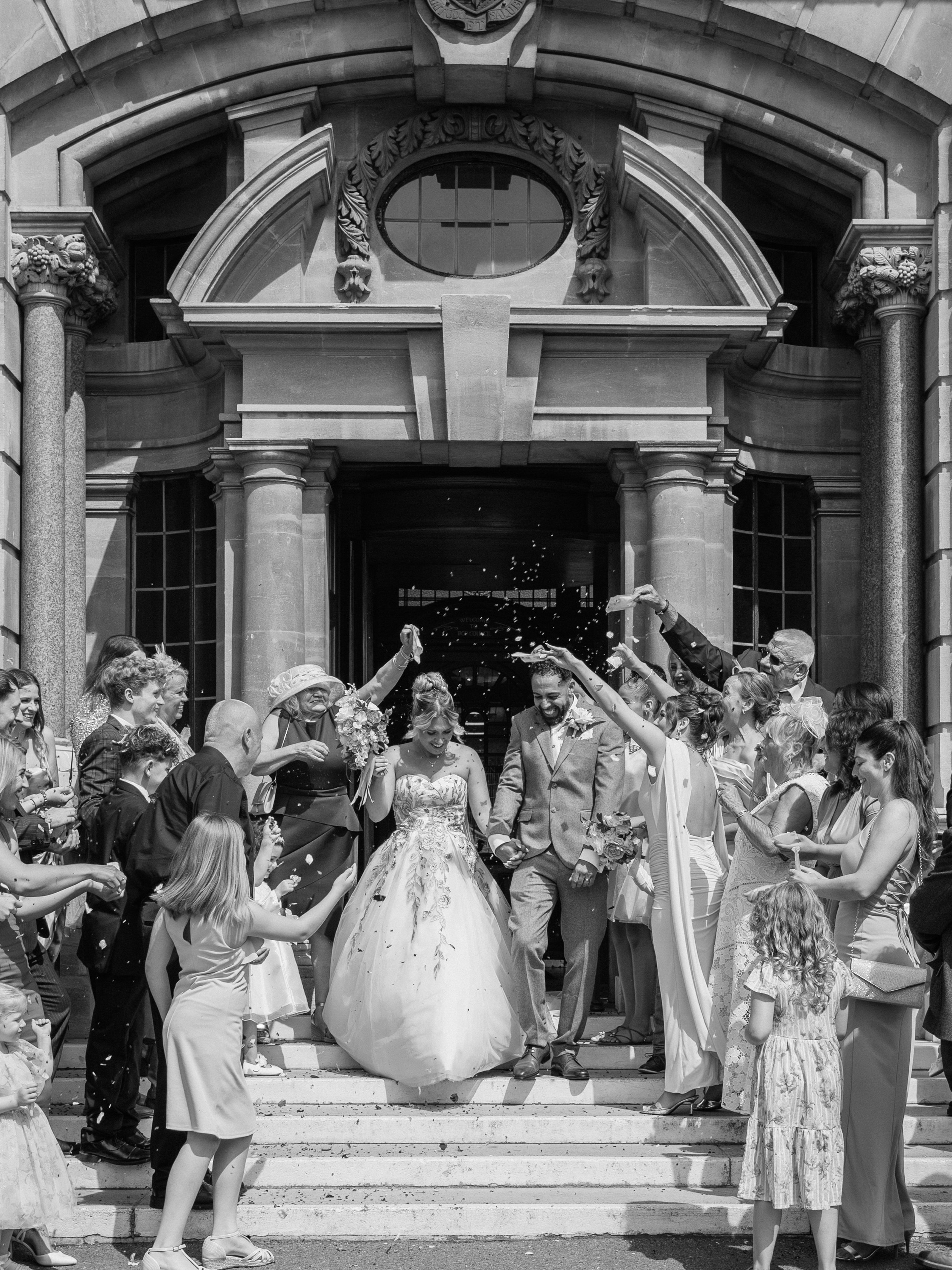 A wedding celebration taking place on the steps of a historic building with columns and decorative stonework. The bride and groom are walking down the steps while guests throw flower petals and applaud.