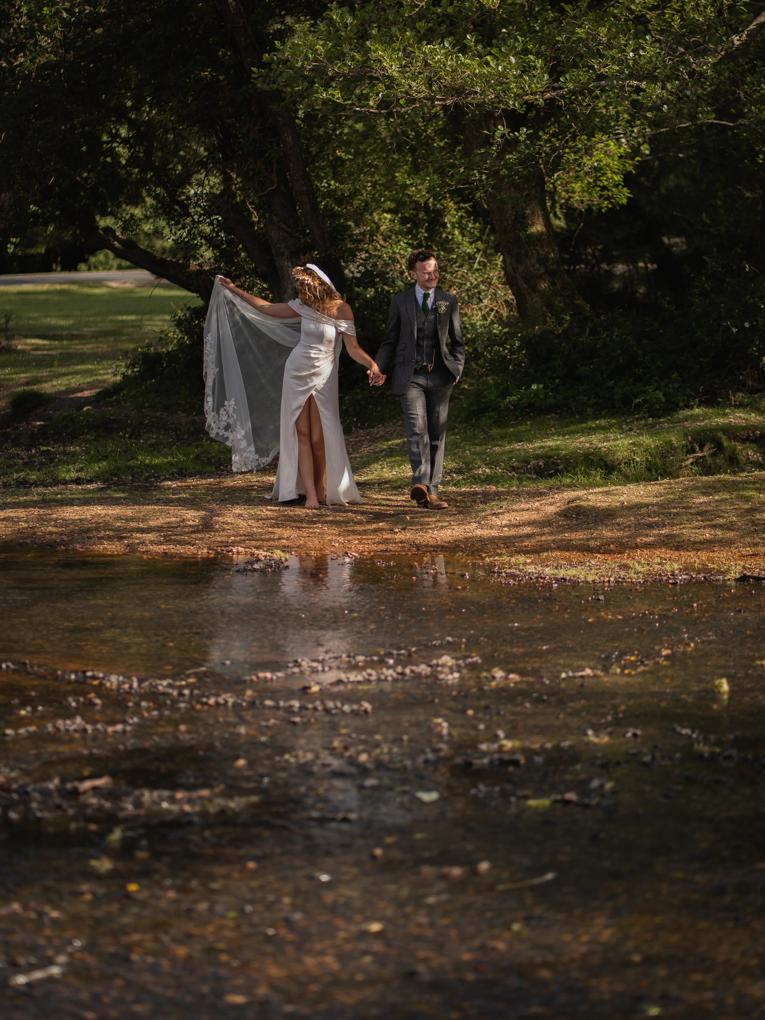 A bride and groom walking hand in hand outdoors, amidst trees with lush green foliage. The bride is dressed in a white wedding gown with a veil, lifting it slightly while walking barefoot. The groom is in a dark suit, smiling and looking to his right