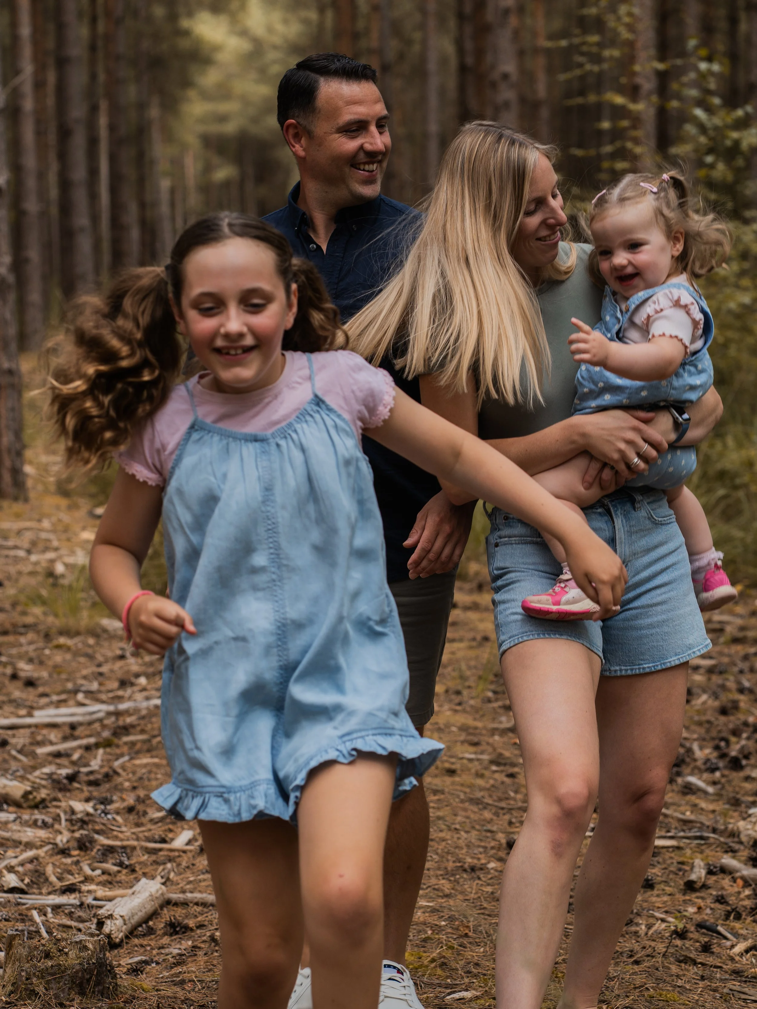 A family of four walking through a forest. The father is smiling, the mother is holding a young girl, and an older girl is in the foreground with her hair in pigtails.