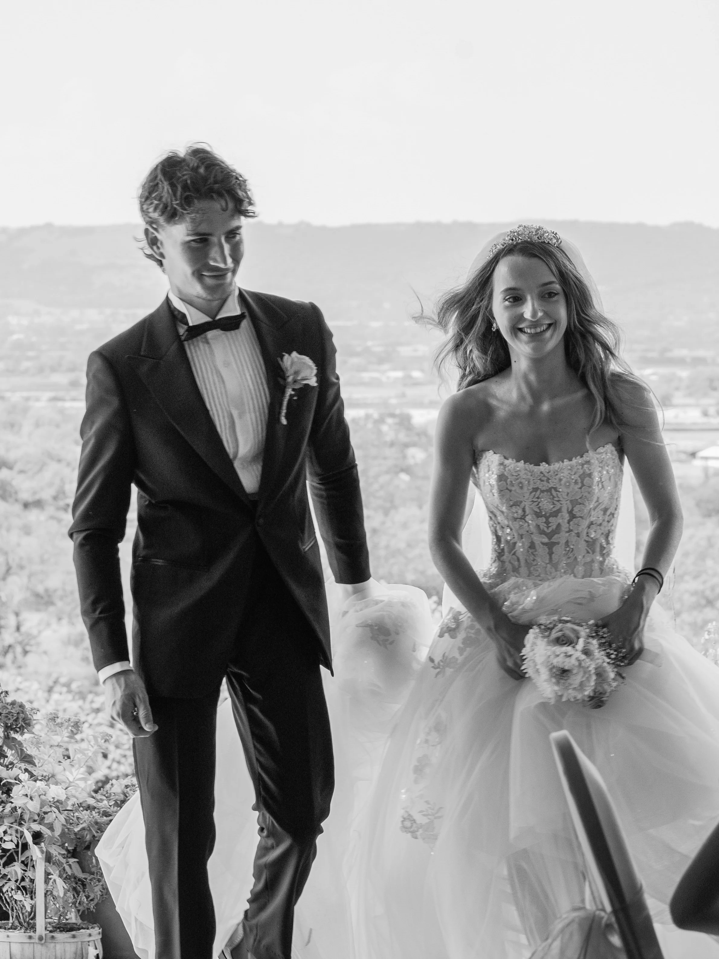 Black and white photo of a bride and groom dressed in wedding attire, smiling and holding hands outdoors with scenic landscape in background.
