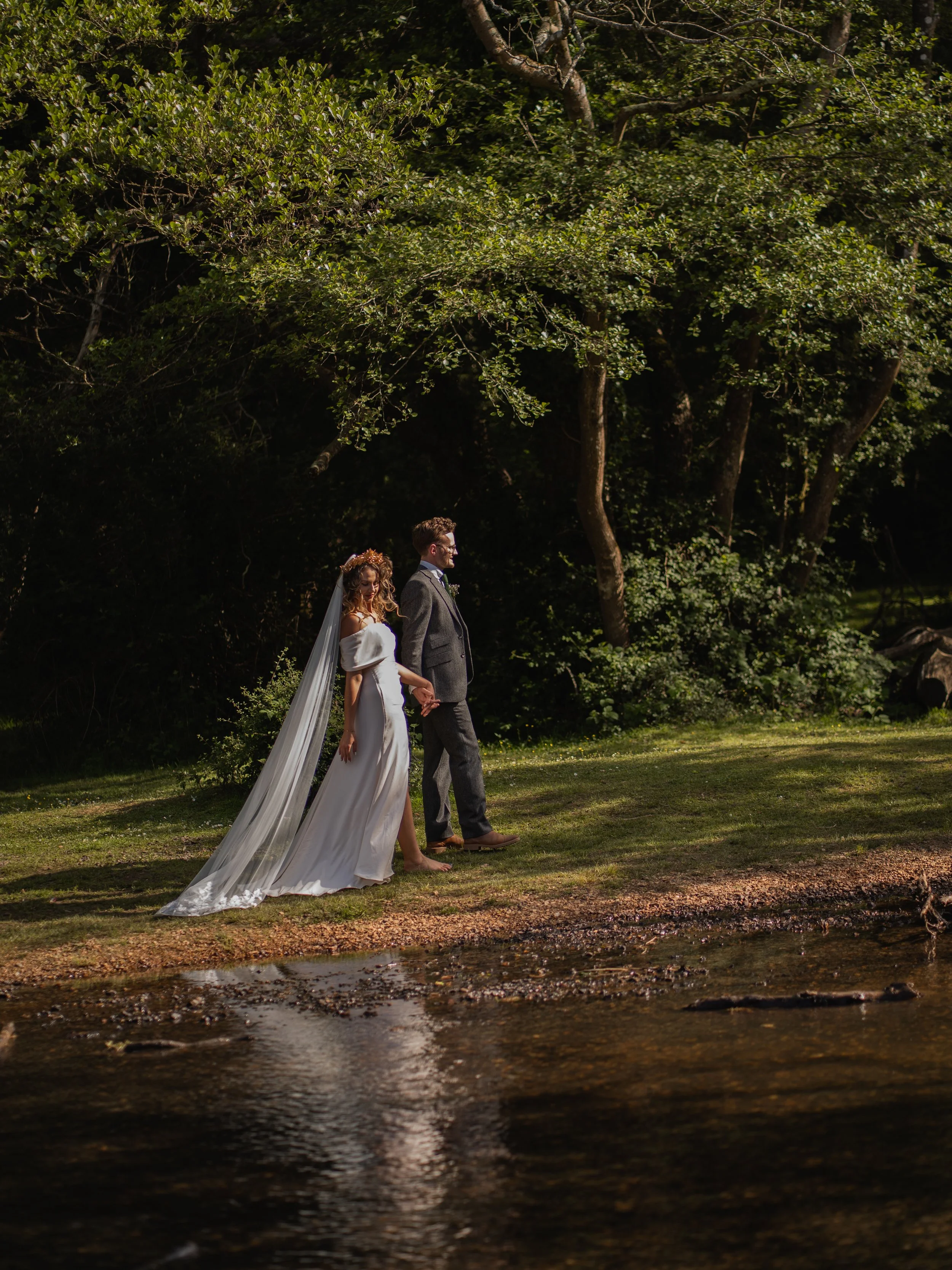A bride and groom holding hands, walking barefoot along a forest stream in daylight, with lush green trees in the background.