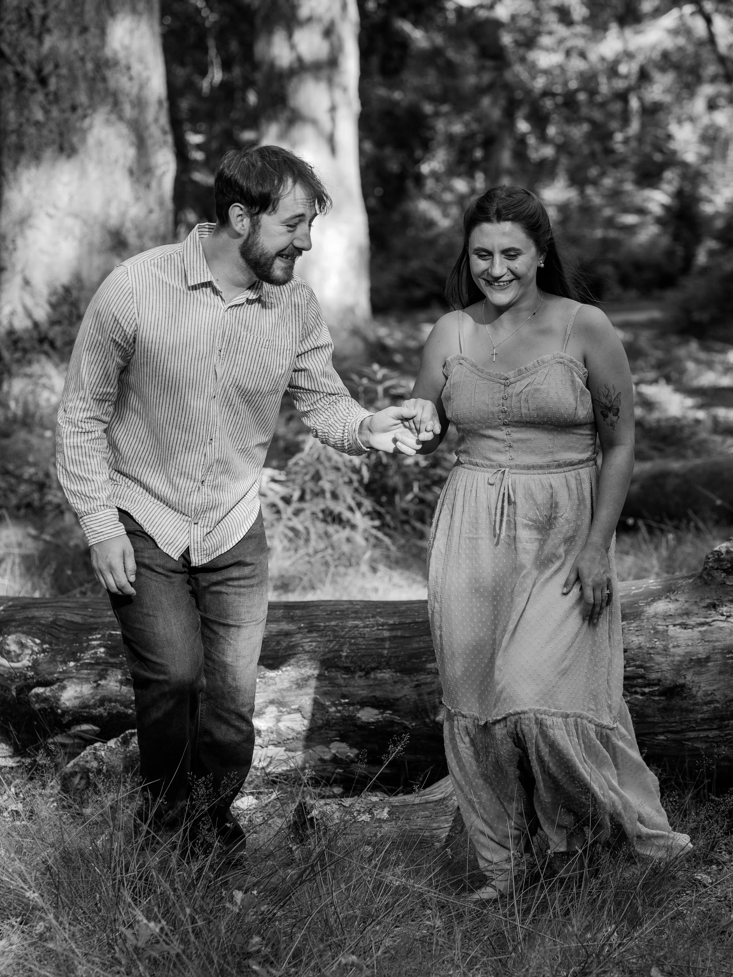 A black and white photo of a man and woman holding hands and smiling, walking through a forested area with tall trees and grass.