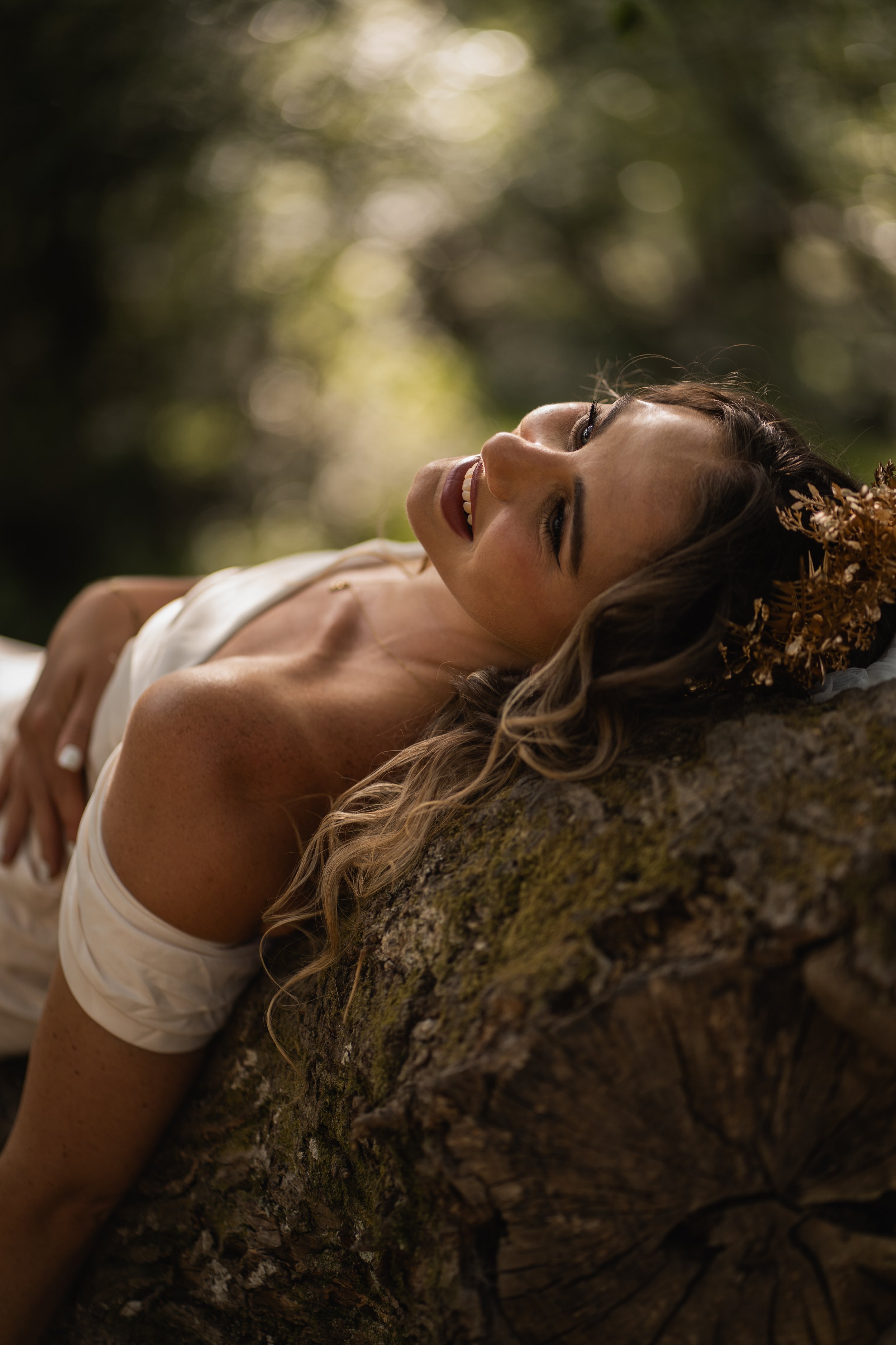 A woman lying on a large tree branch, smiling and looking up, with a blurred background of sunlight filtering through leaves, wearing a white top and a floral headpiece.