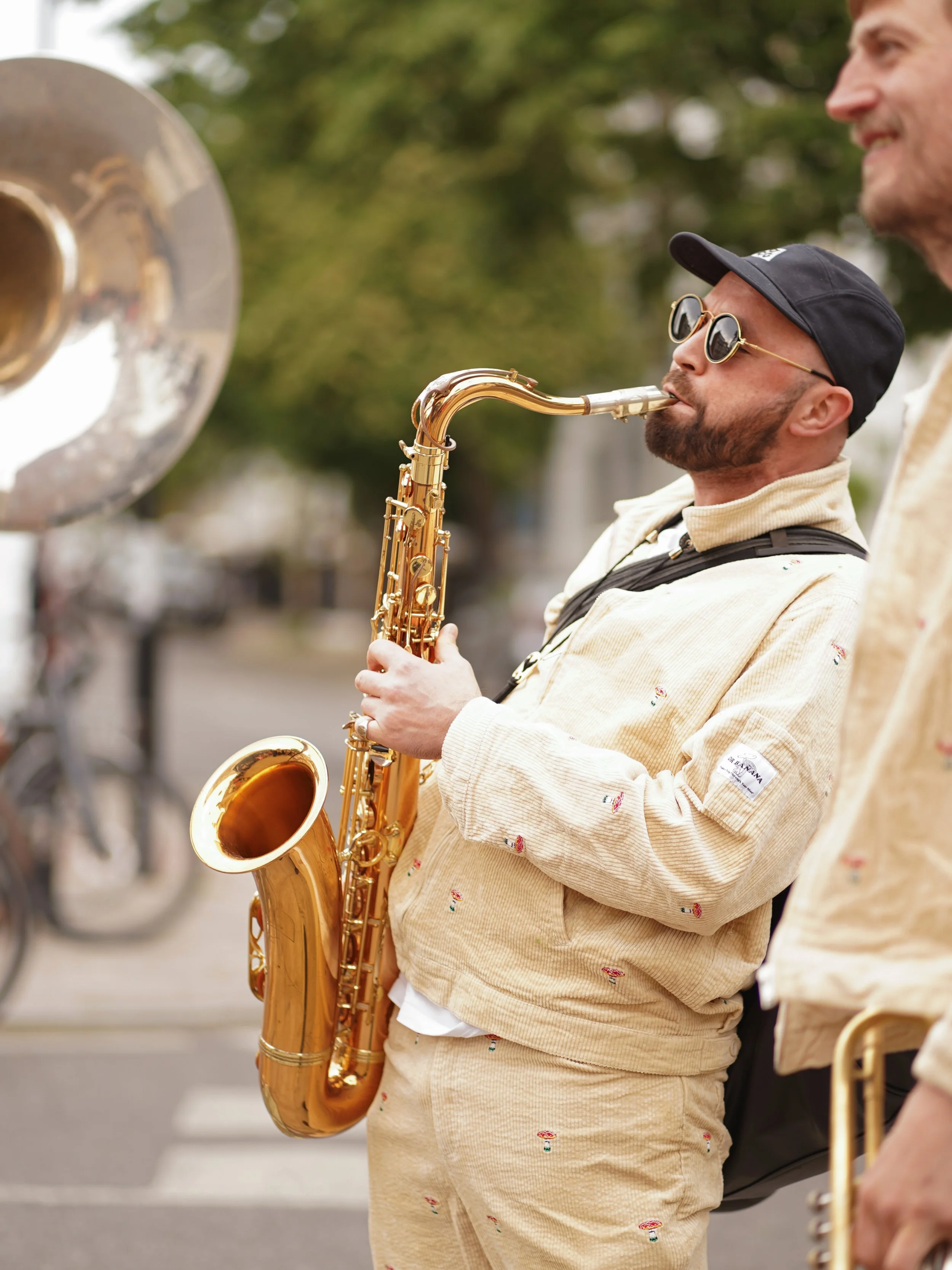 A man playing a saxophone at an outdoor event, with another person standing beside him. The background shows blurred greenery and a street.