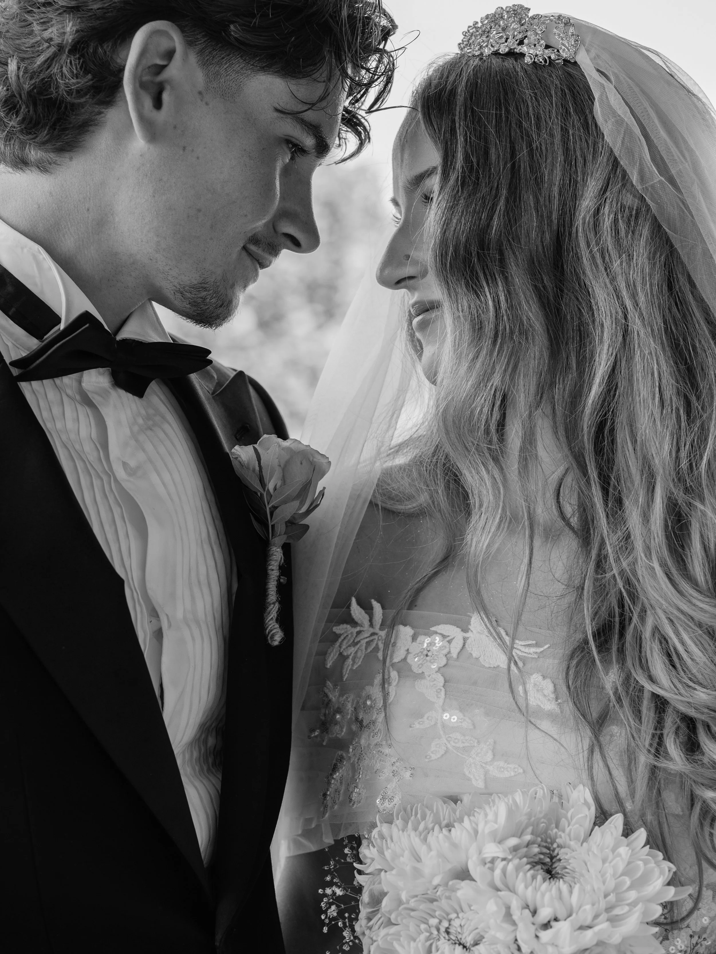 Black and white close-up of a bride and groom with their foreheads touching, on their wedding day. The groom wears a tuxedo with a bow tie, and the bride has a veil and a tiara, holding a bouquet of large flowers.