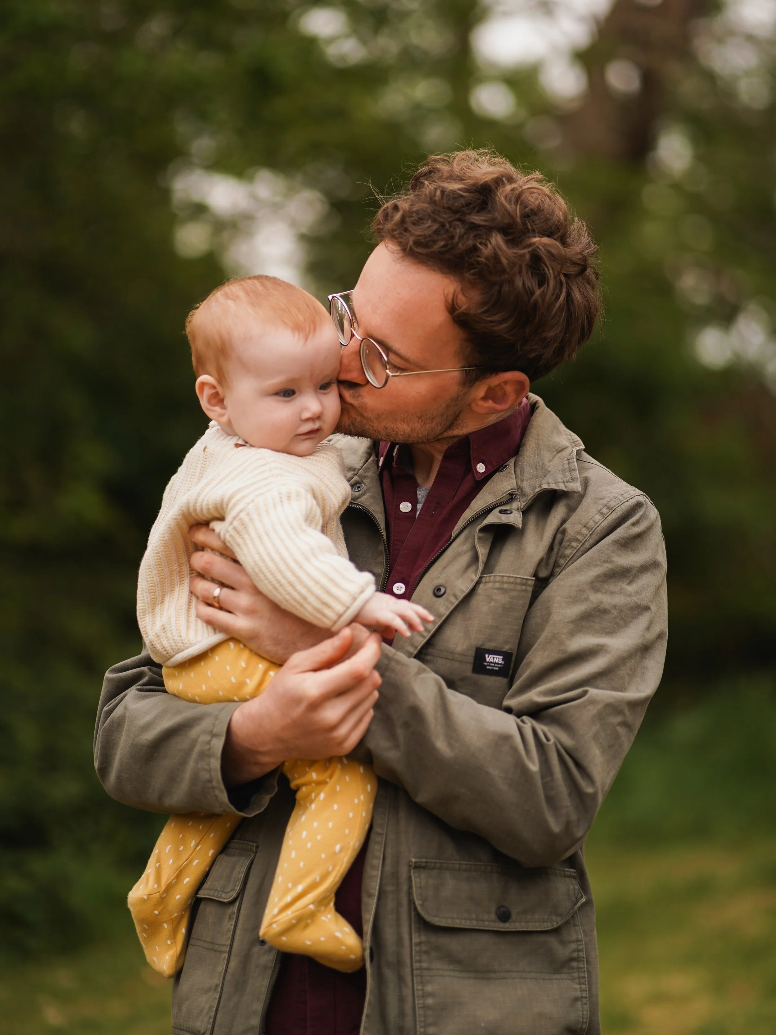 A man with curly brown hair and glasses holding a toddler with red hair, in an outdoor setting with blurred green trees and foliage in the background. The man is kissing the child's cheek.