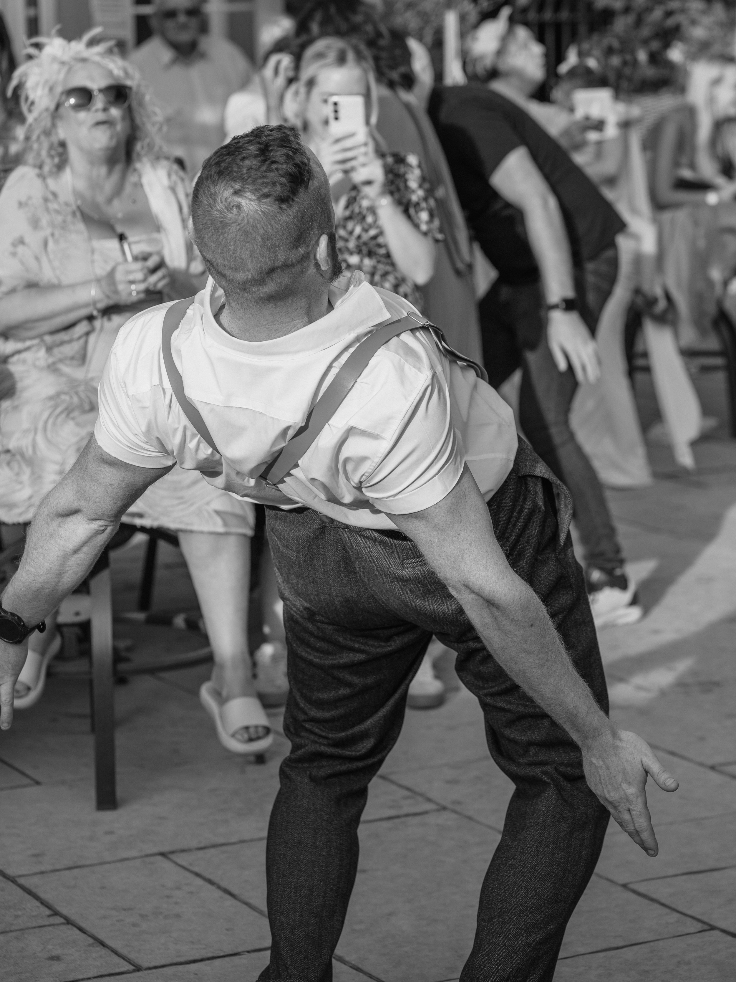 A man in formal attire performing a dance move outdoors, with an audience of women watching and taking photos, some sitting on chairs and others standing.
