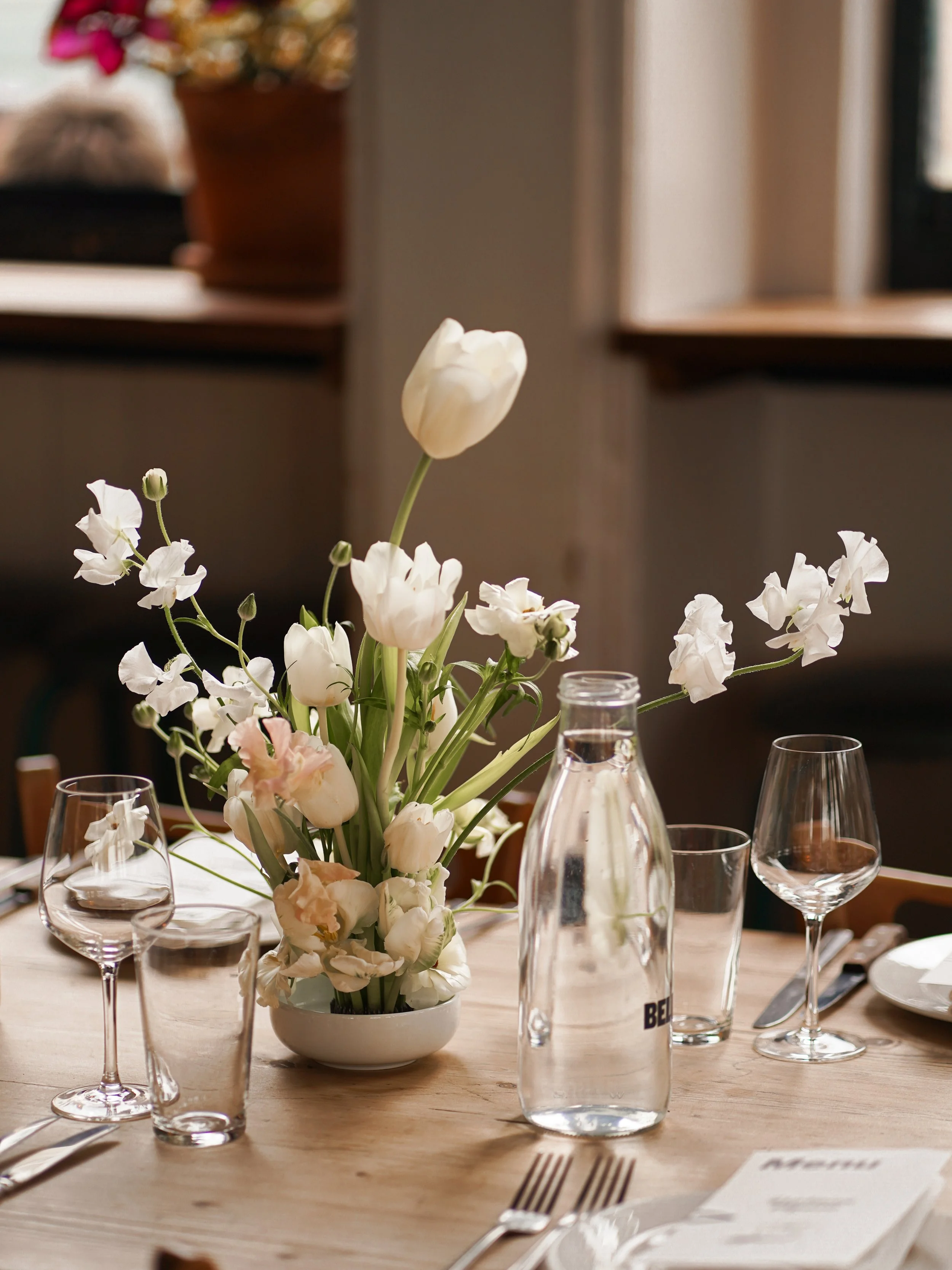 A table decorated with a floral arrangement of white tulips and other white flowers, set with wine glasses, water bottles, utensils, and a printed menu, in a cozy indoor setting.