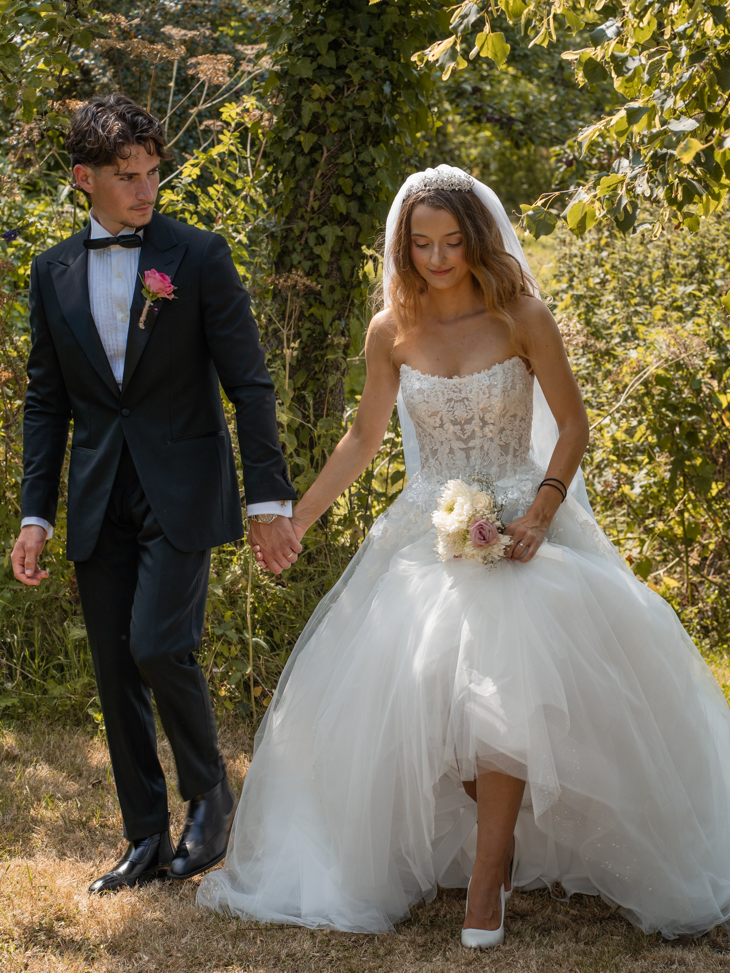 A bride and groom walking hand in hand outdoors, surrounded by greenery.