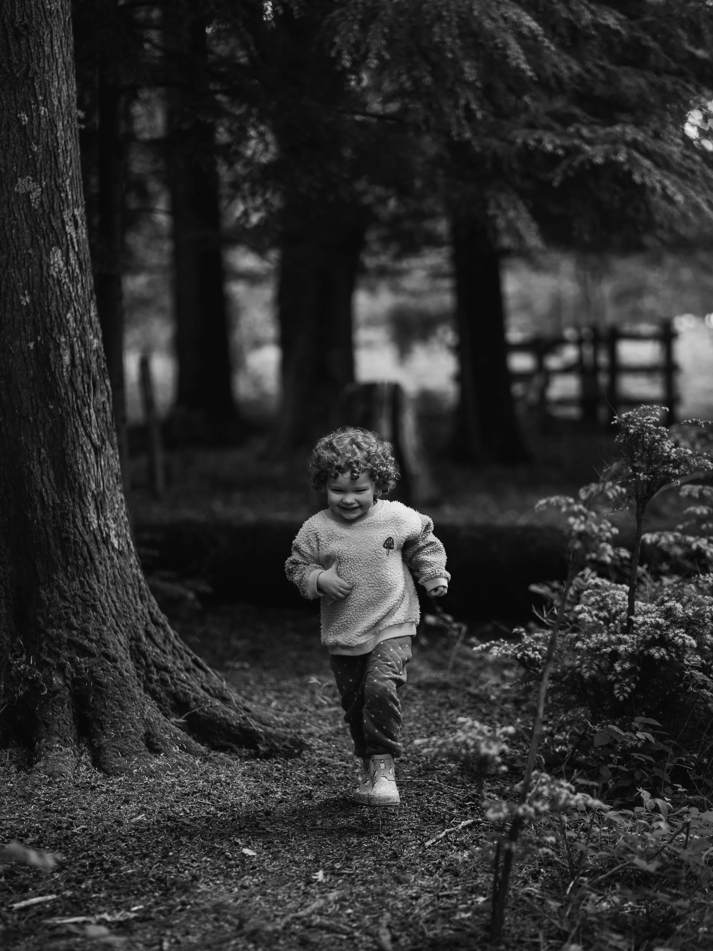 A child with curly hair running happily in a forest, smiling, wearing a fluffy sweater and patterned pants, with trees in the background in Wilverly in Brockenhurst in the New Forest for a family photoshoot.