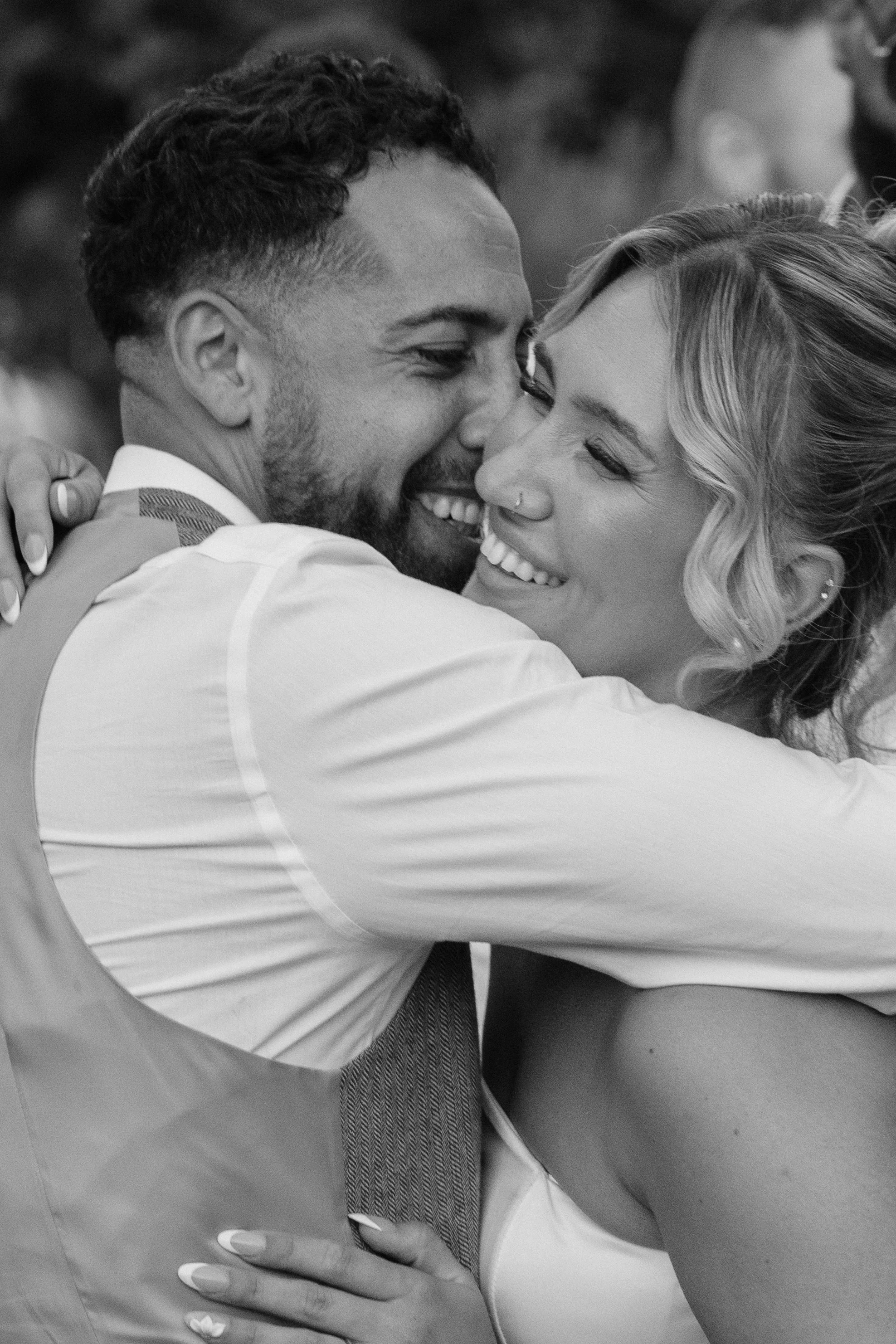 A black and white photo of a smiling man and woman embracing each other closely, with their faces touching. The man has short, curly hair and a beard, and is wearing a dress shirt. The woman has wavy, shoulder-length hair, a nose ring, and is dressed
