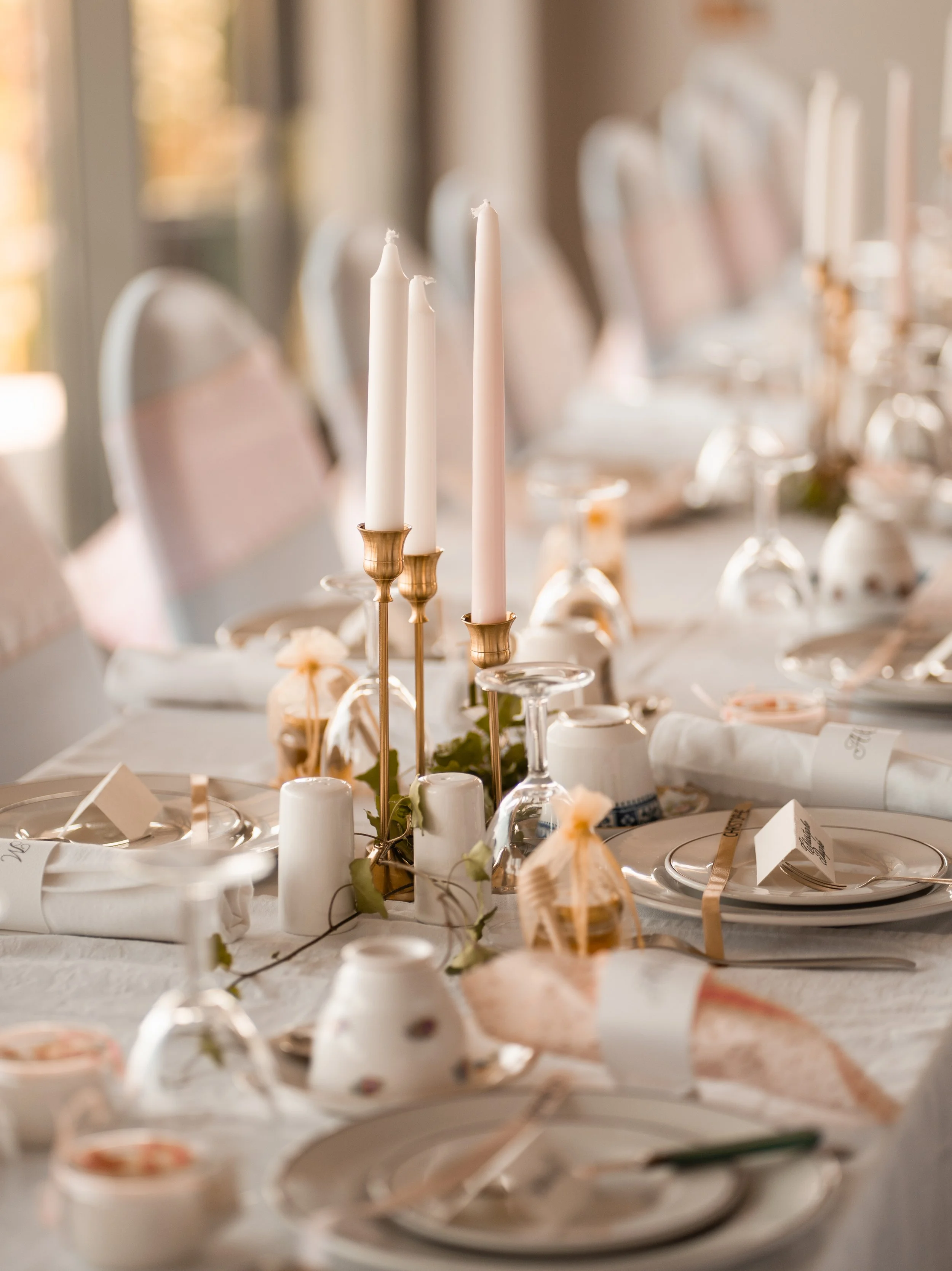 Elegant banquet table set with white candles in gold candleholders, white plates, silverware, wine glasses, and small decorative items, with a soft, warm lighting ambiance.