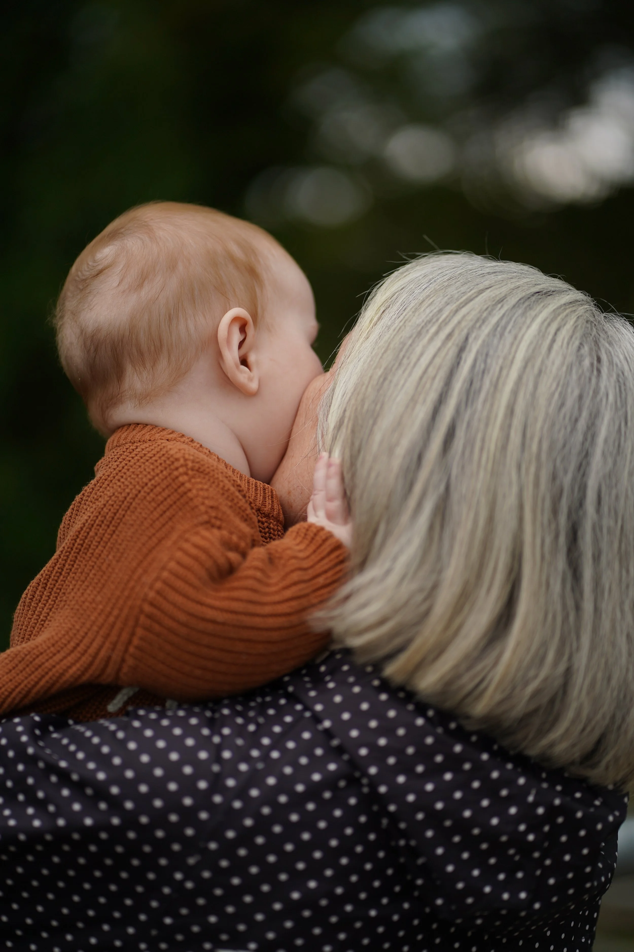 Close-up of a young child with red hair, wearing a brown sweater, being held close by an elderly woman with long, white hair, wearing a dark polka dot shirt, outdoors with blurred greenery background.