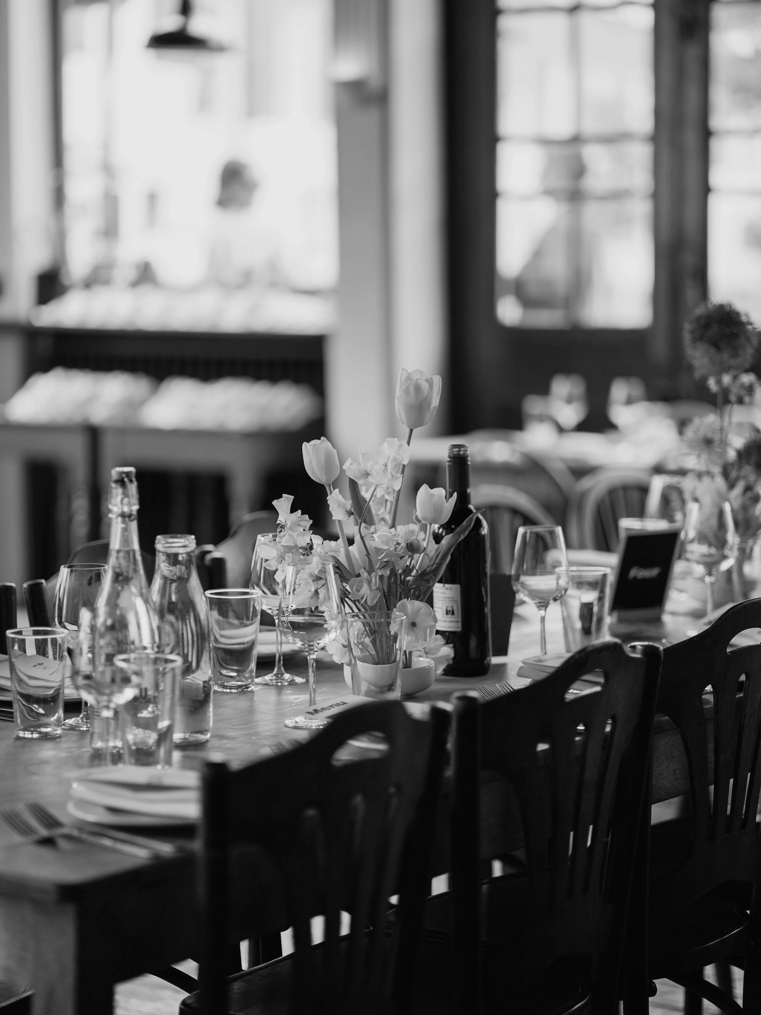 A black and white photo of a dining table set with glasses, bottles, and a floral centerpiece in a well-lit restaurant or café with large windows and wooden chairs.
