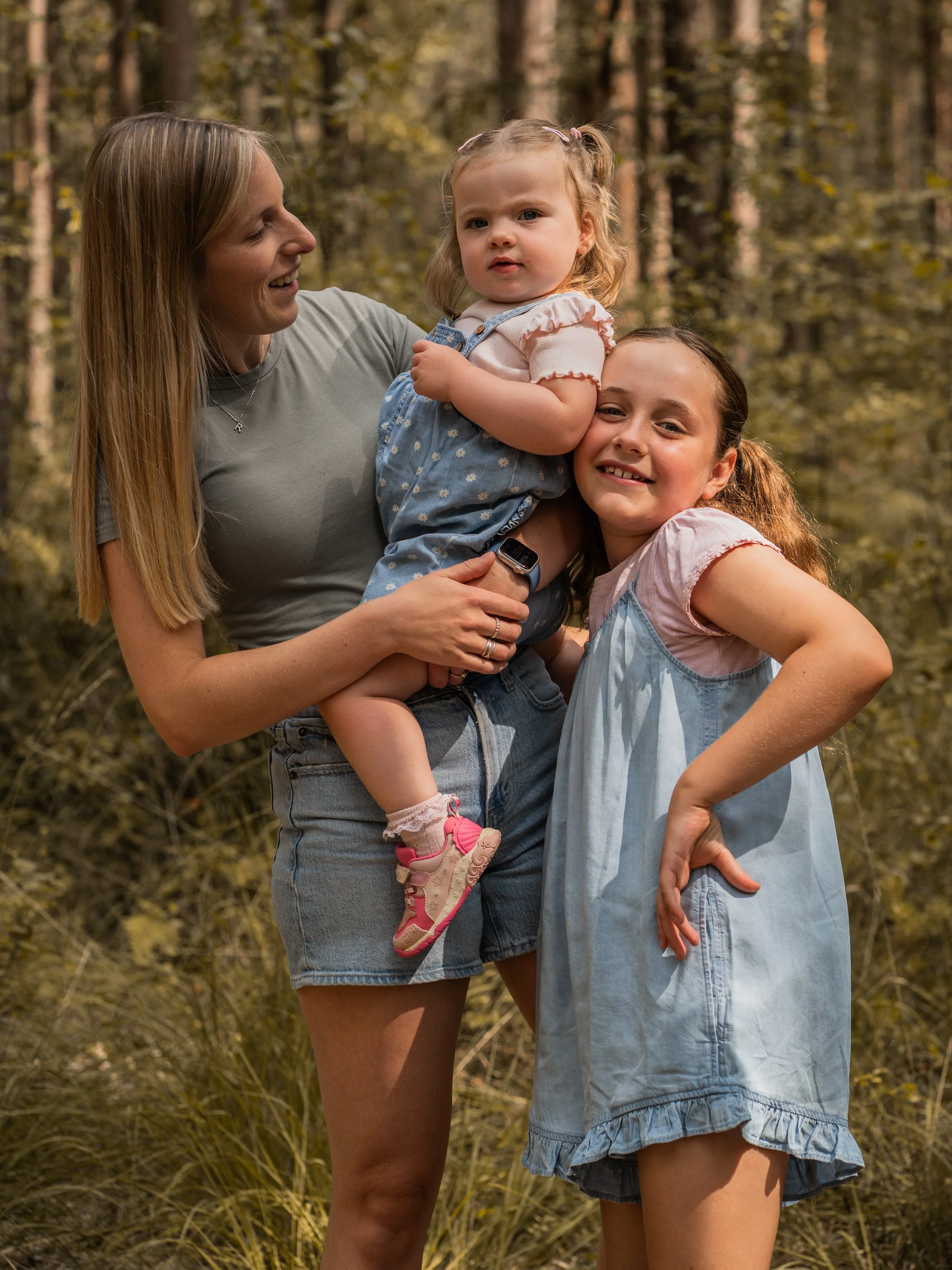 A woman with long blonde hair smiling and holding a young girl with blonde hair and pigtails in a forest setting. The girl has a serious expression and is wearing a light pink top with ruffled sleeves and denim overalls. Another girl with long brown 