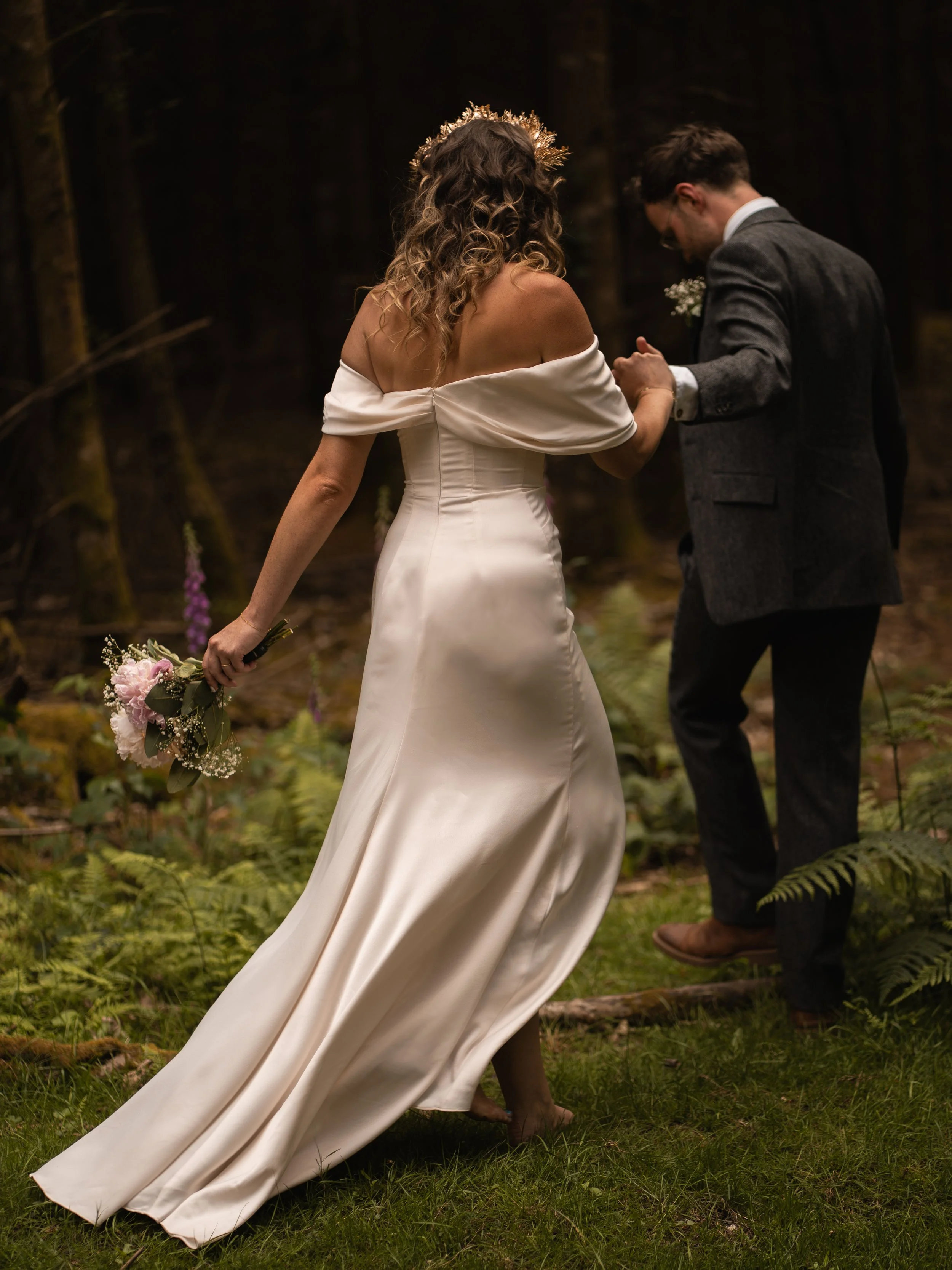 Bride and groom holding hands in a forest during their wedding ceremony, with the bride wearing an off-the-shoulder white gown and holding a pink flower bouquet.