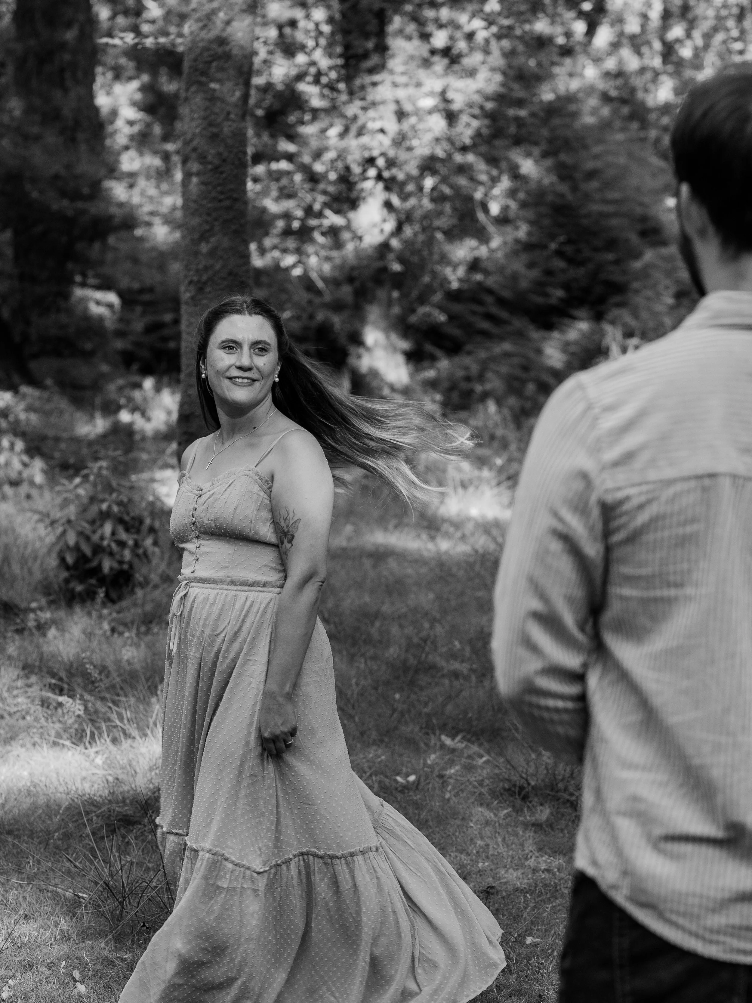 A woman in a sleeveless dress smiling at a man in a striped shirt outdoors in a wooded area.