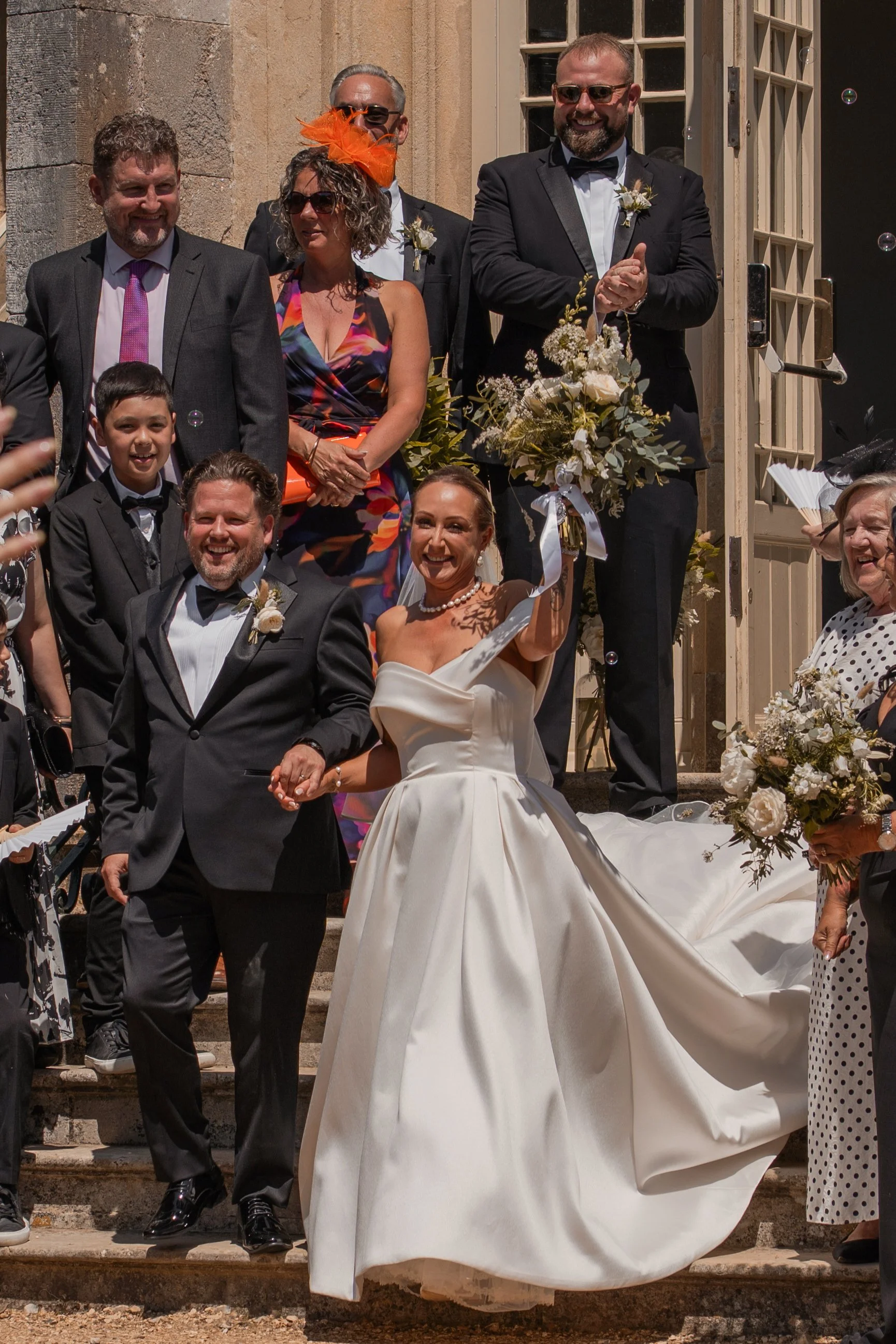 Wedding group photo outside a stone building with stairs, featuring groom and bride in the center, surrounded by family and friends dressed in formal attire, with some wearing sunglasses and holding bouquets.