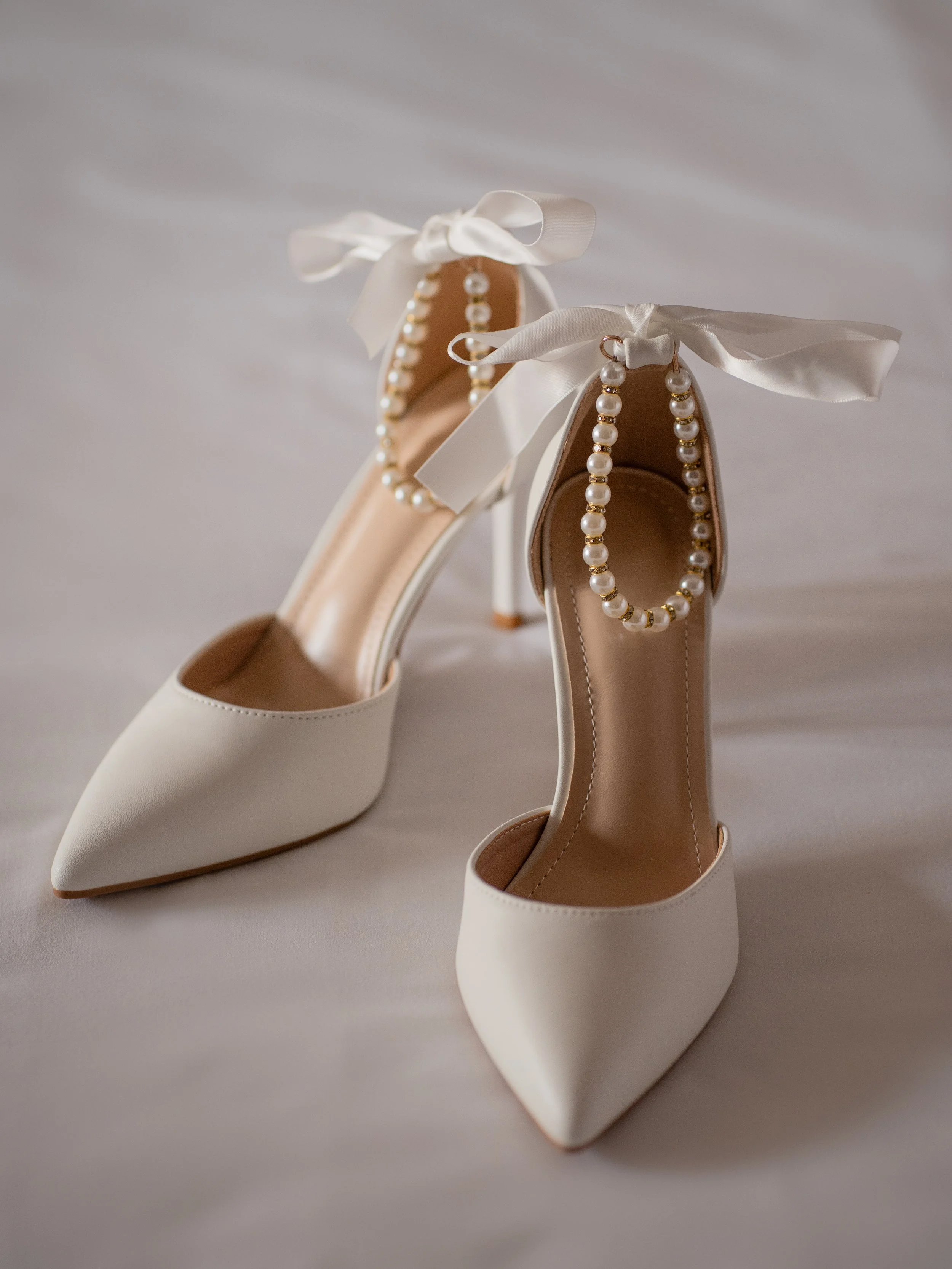 Pair of white high-heeled shoes decorated with pearl ankle straps and satin ribbons tied in bows.