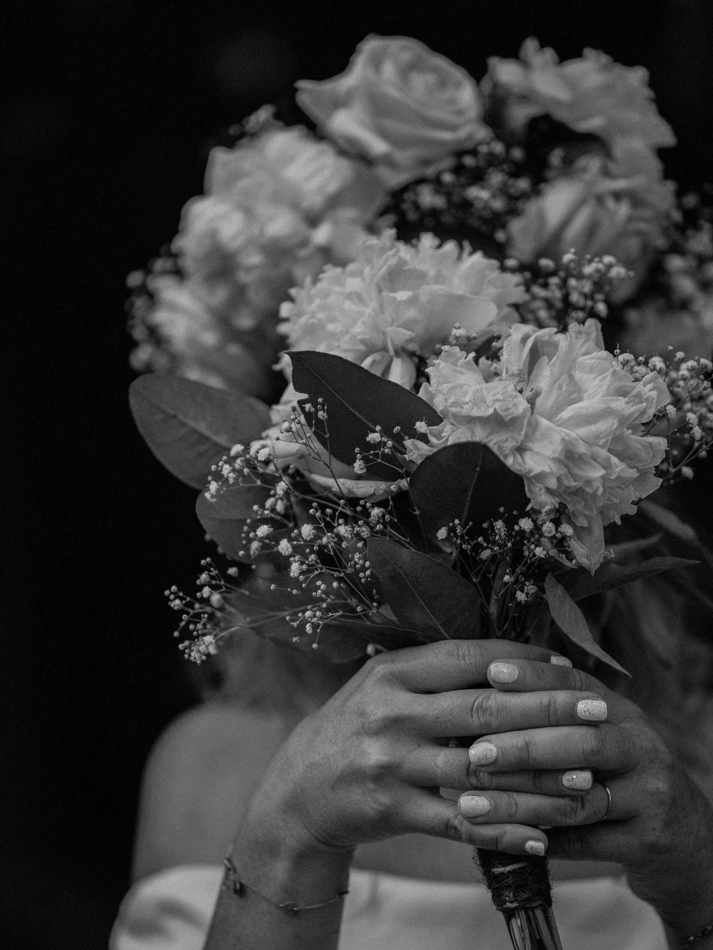A black and white photo of a person’s hand holding a bouquet of flowers, including roses and small blossoms, with the person's other hand wrapped around it. The person has painted nails and is wearing a ring and bracelet.