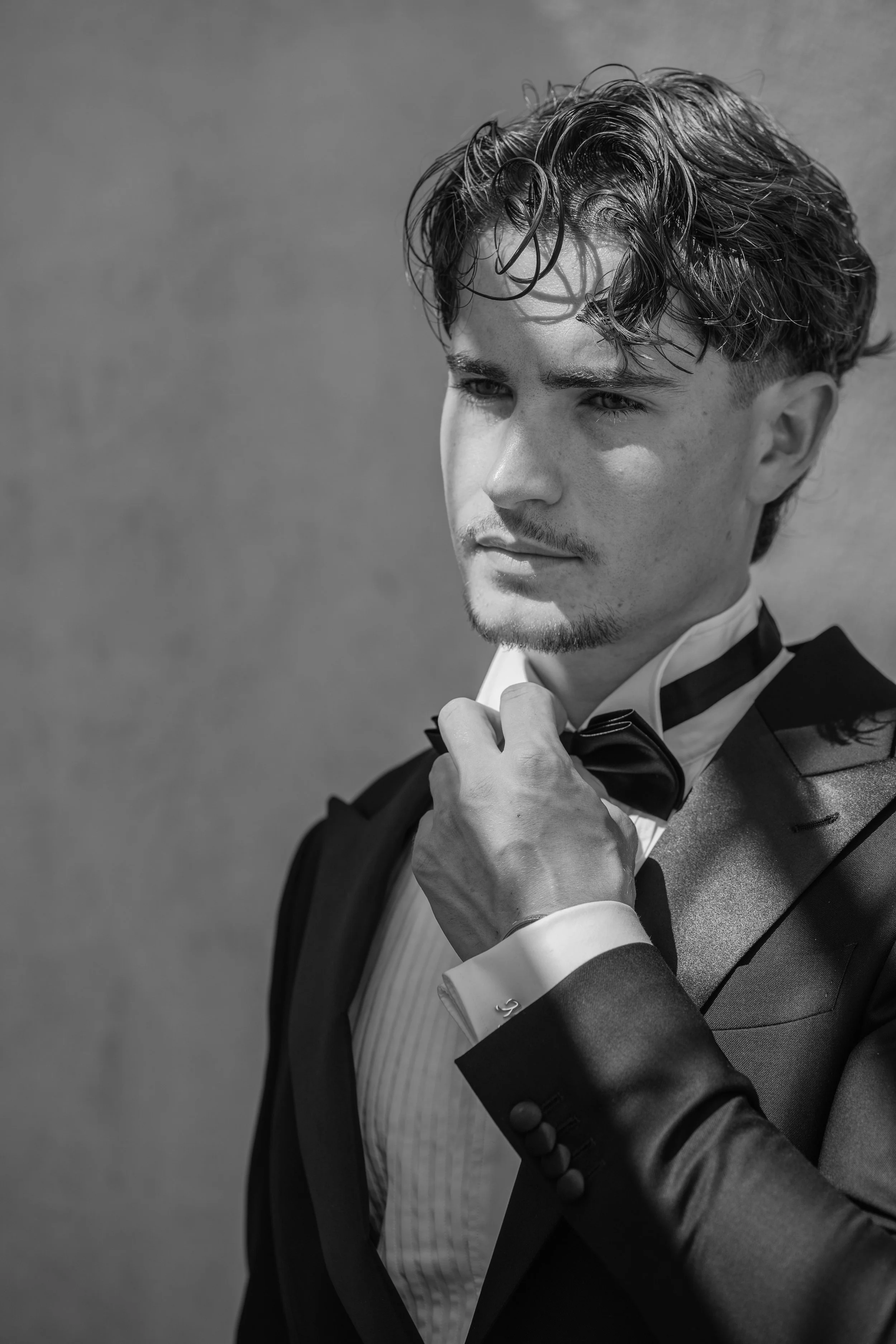 A black and white photo of a young man in a tuxedo adjusting his bow tie, looking thoughtfully to the side.