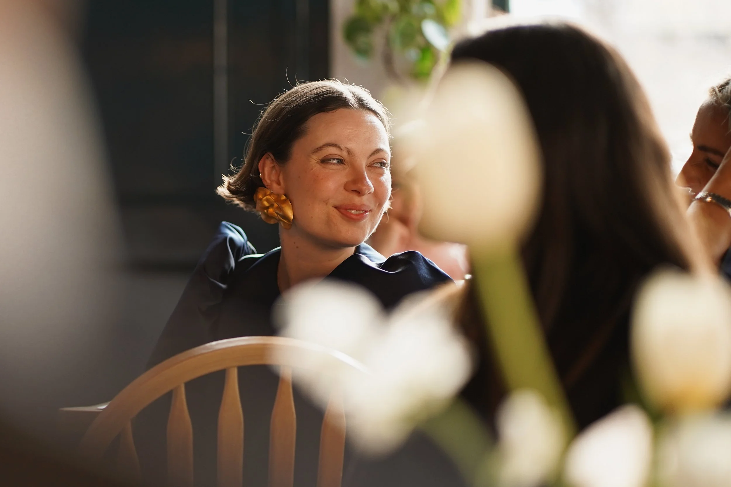 A woman with short brown hair, wearing gold earrings and a dark-colored top, smiling and talking to someone. There are other women visible in the background, with a blurred foreground of a green plant and wooden chairs.