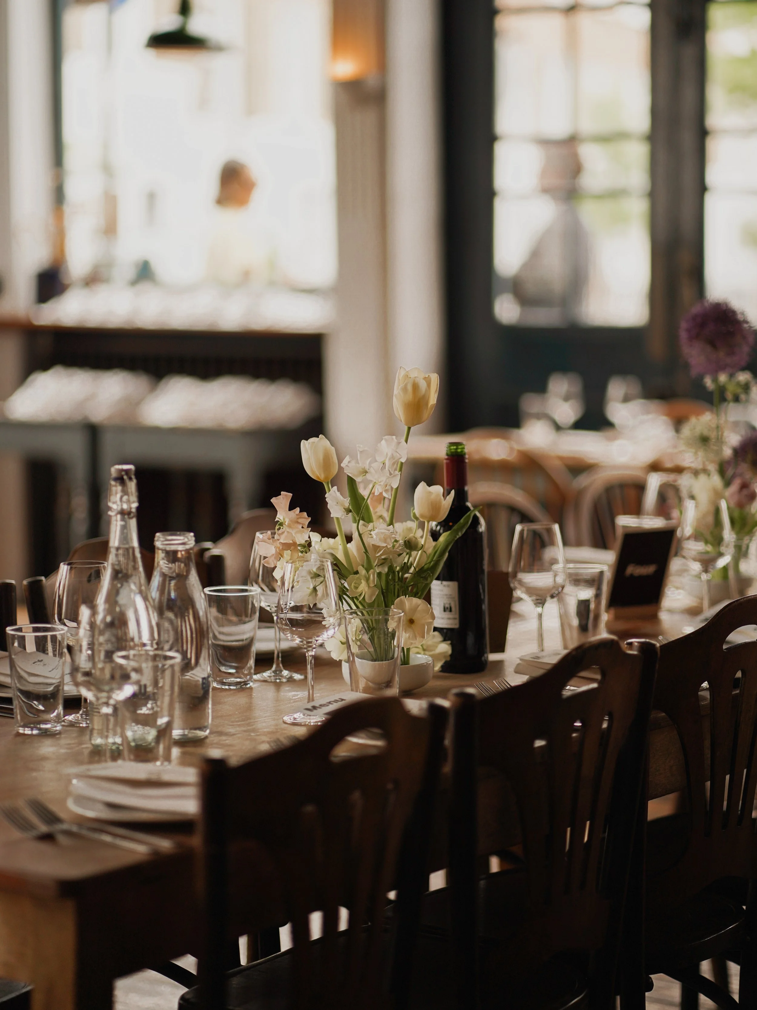 A cozy dining setup with a wooden table decorated with white flowers in a glass vase, surrounded by empty glasses and bottles. The background shows large windows and a blurred figure, indicating an indoor space with natural light.