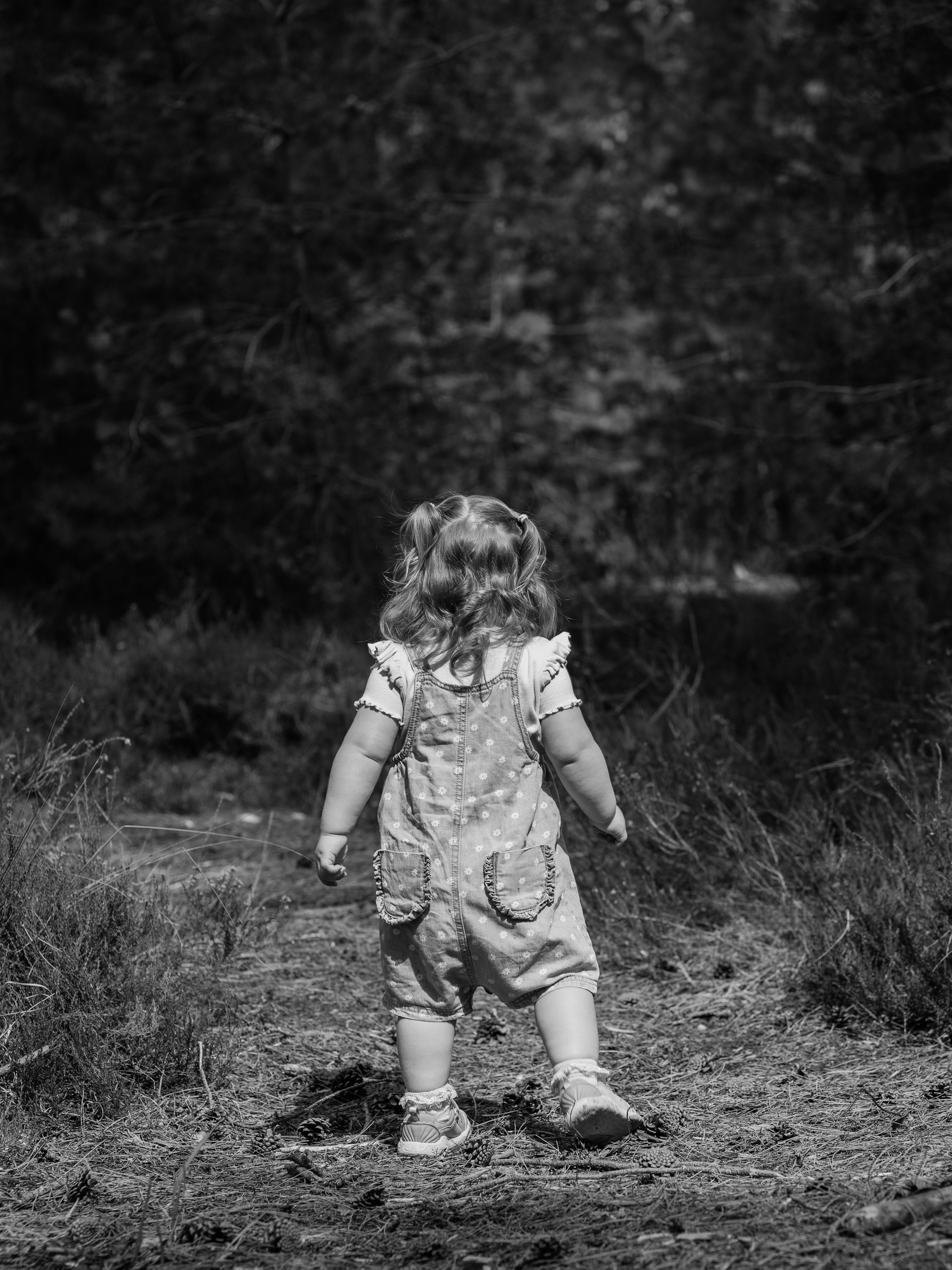 Back view of a young girl with curly hair walking on a dirt path in a natural outdoor setting with trees and grass, captured in black and white.