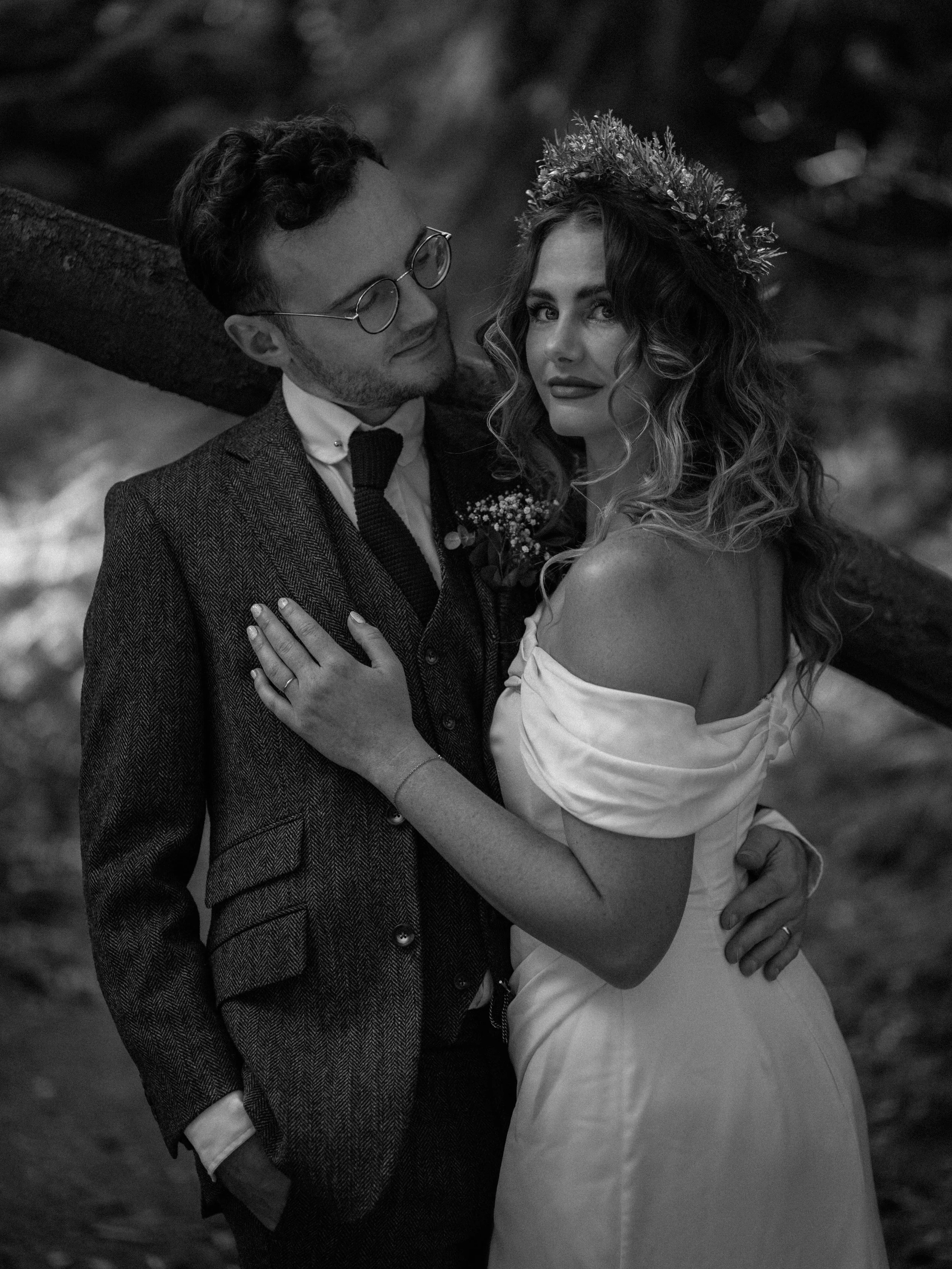 A black and white photo of a man and woman embracing outdoors. The man wears round glasses and a suit; the woman wears a dress and a floral crown. They are looking at each other lovingly.