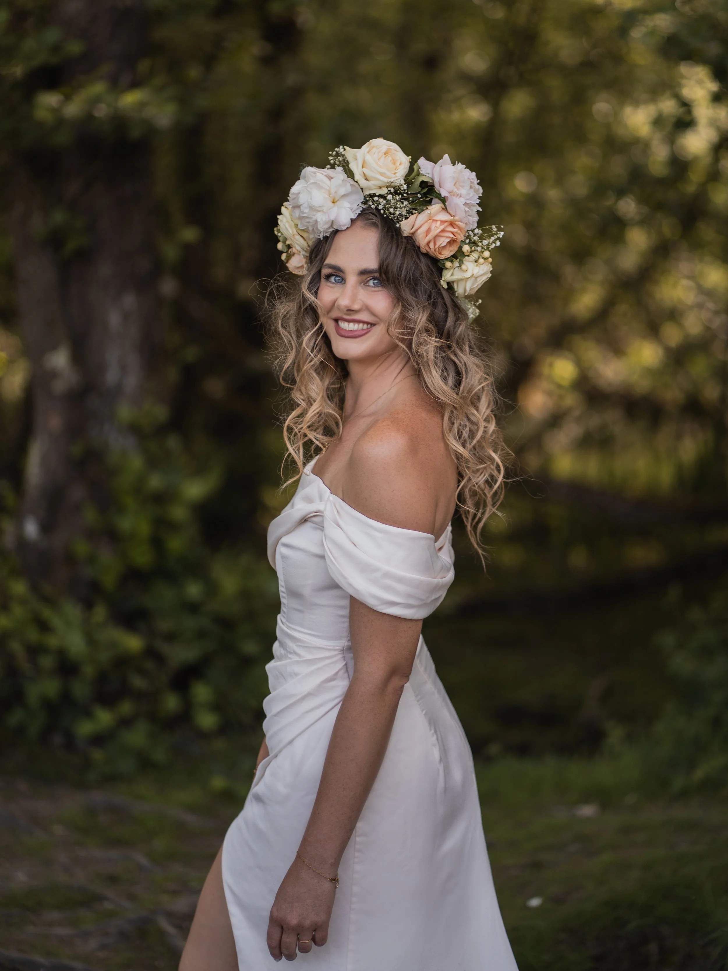 A young woman with curly blonde hair and blue eyes, wearing a white off-the-shoulder dress and a large floral crown of roses and baby's breath, standing outdoors with trees and natural scenery in the background, smiling at the camera.