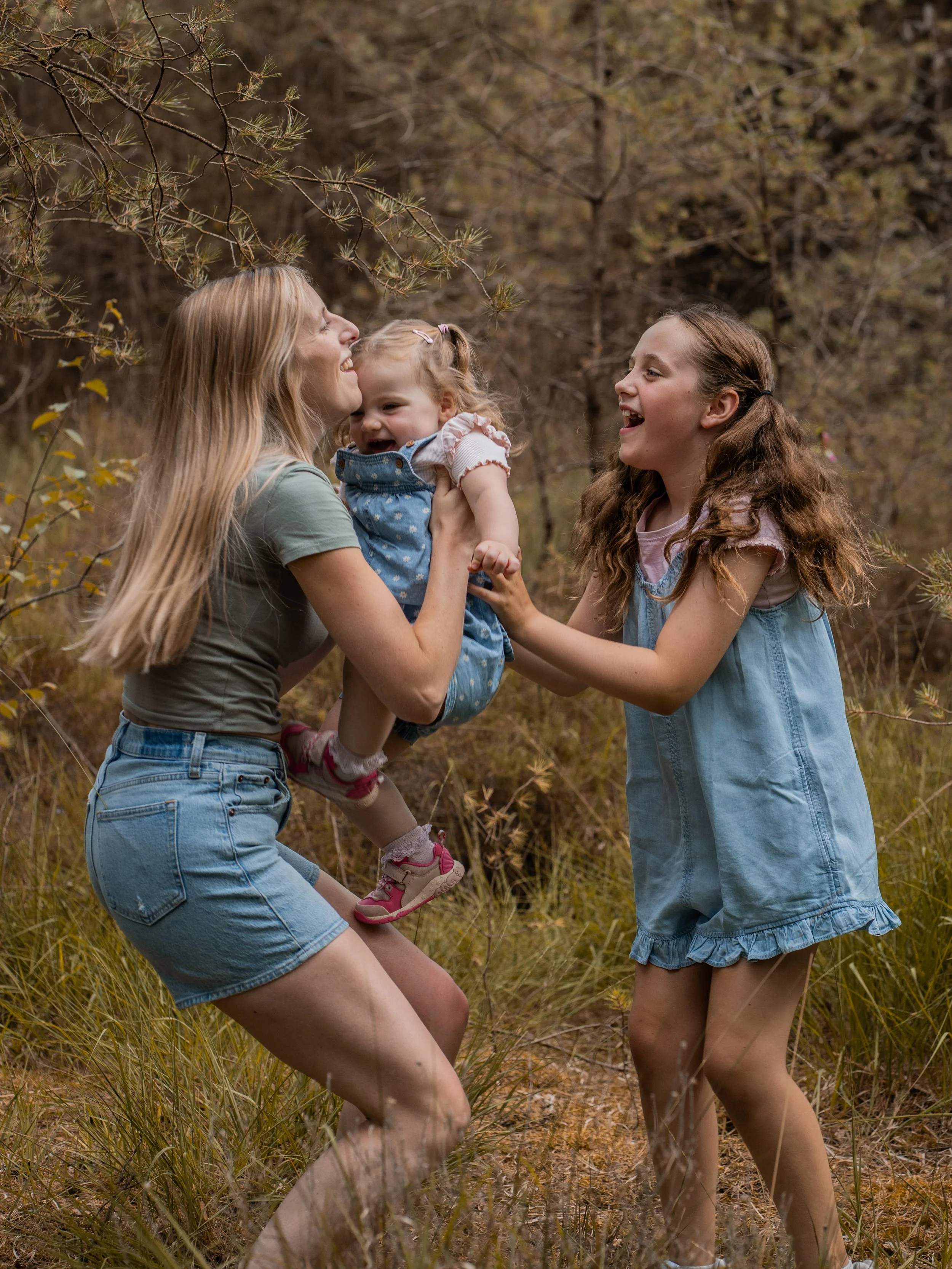 Three people, two women and a young girl, are outdoors in a wooded area, playing and laughing together.