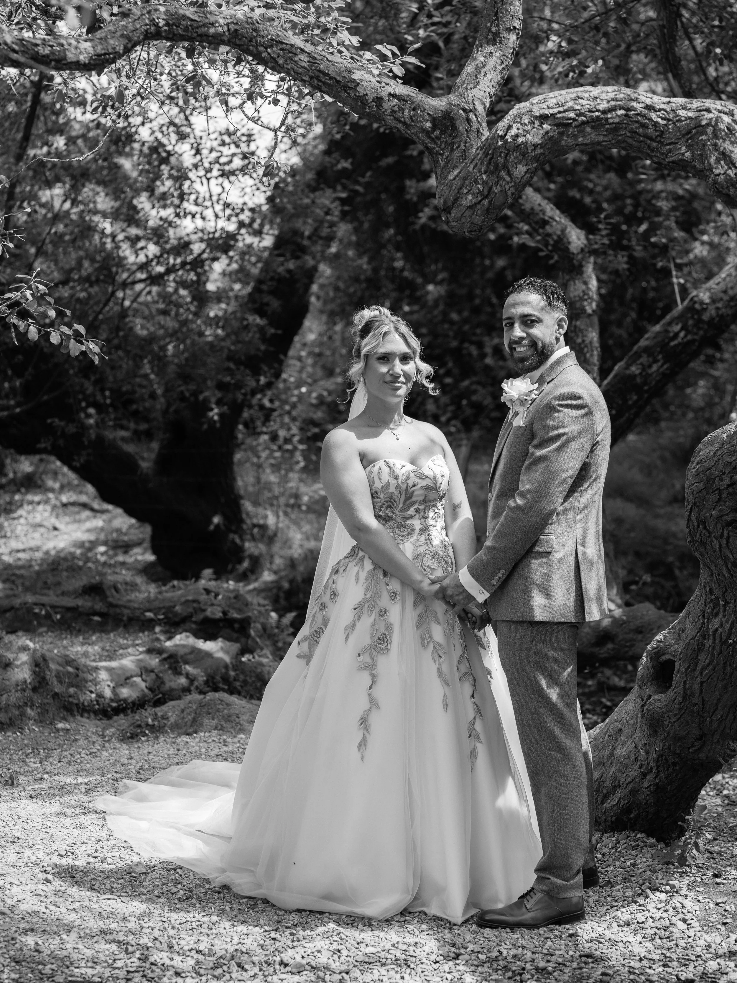 A black-and-white photo of a bride and groom standing outdoors in a natural setting with trees. The bride is wearing a strapless wedding gown with a floral pattern and a veil, while the groom is wearing a suit with a boutonniere. They are holding han
