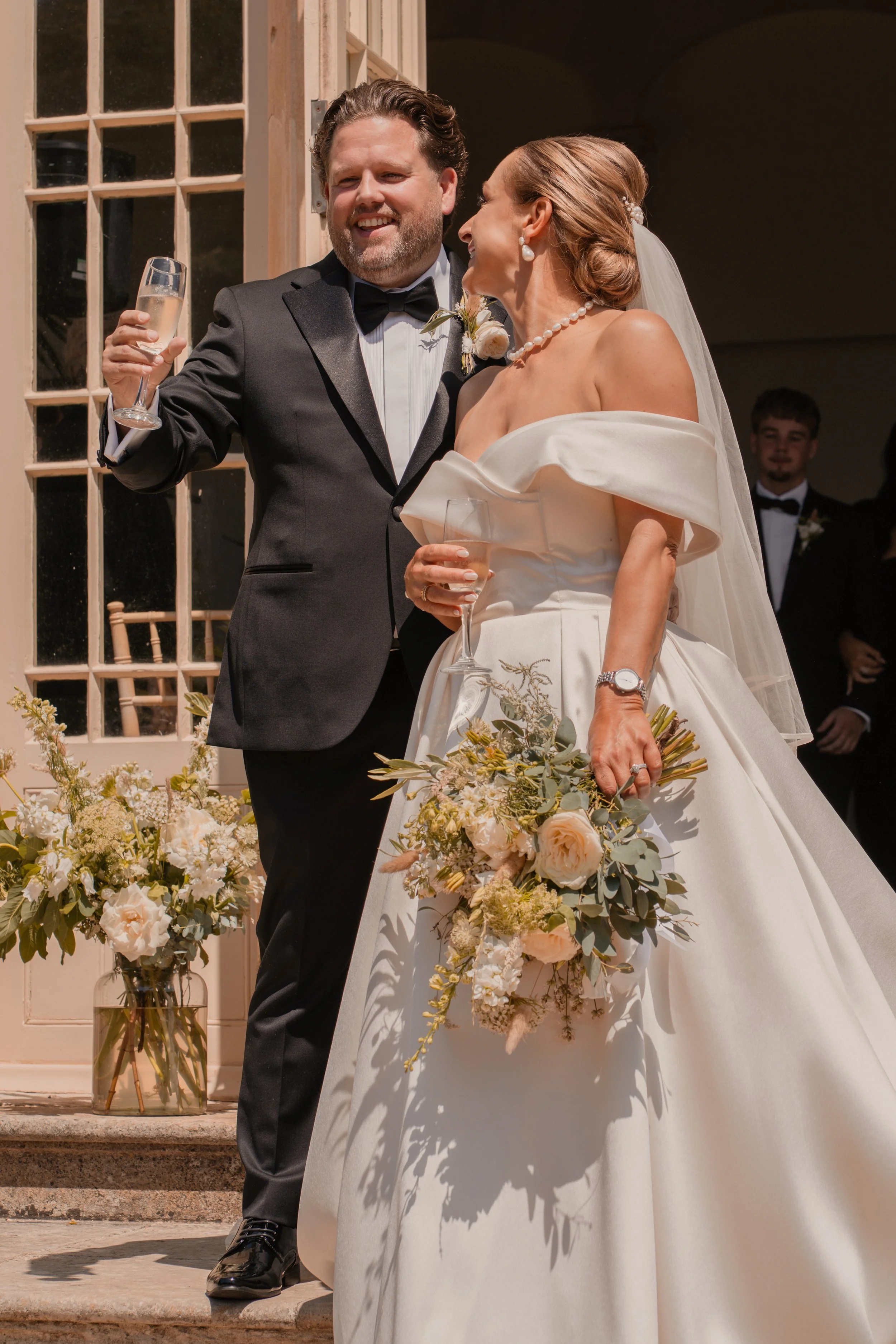 A bride and groom celebrating at their wedding, holding glasses of champagne, with floral arrangements in front of a wooden door.