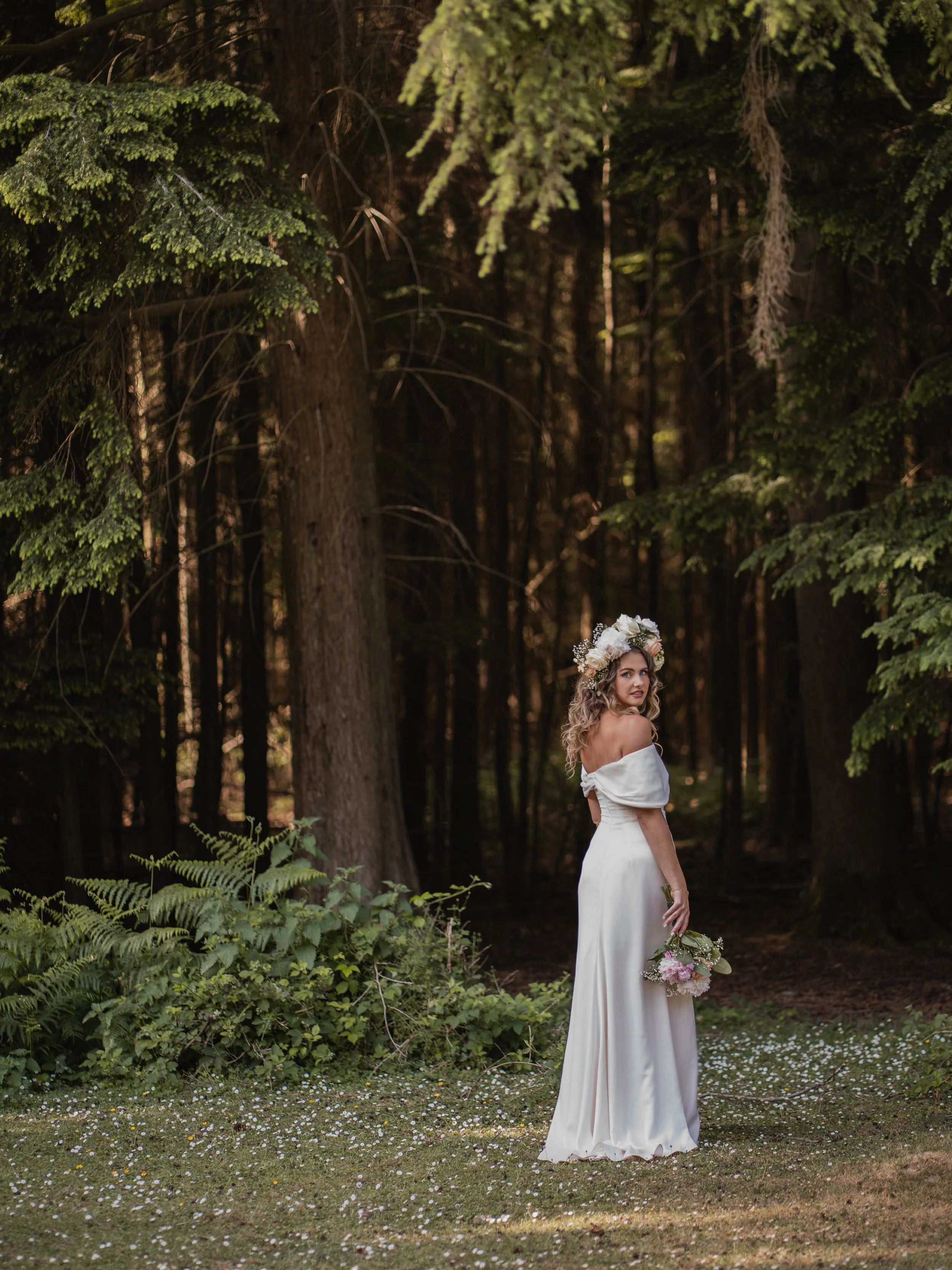 A woman in a white wedding dress with a floral crown holding a bouquet stands in a wooded area surrounded by trees and greenery.