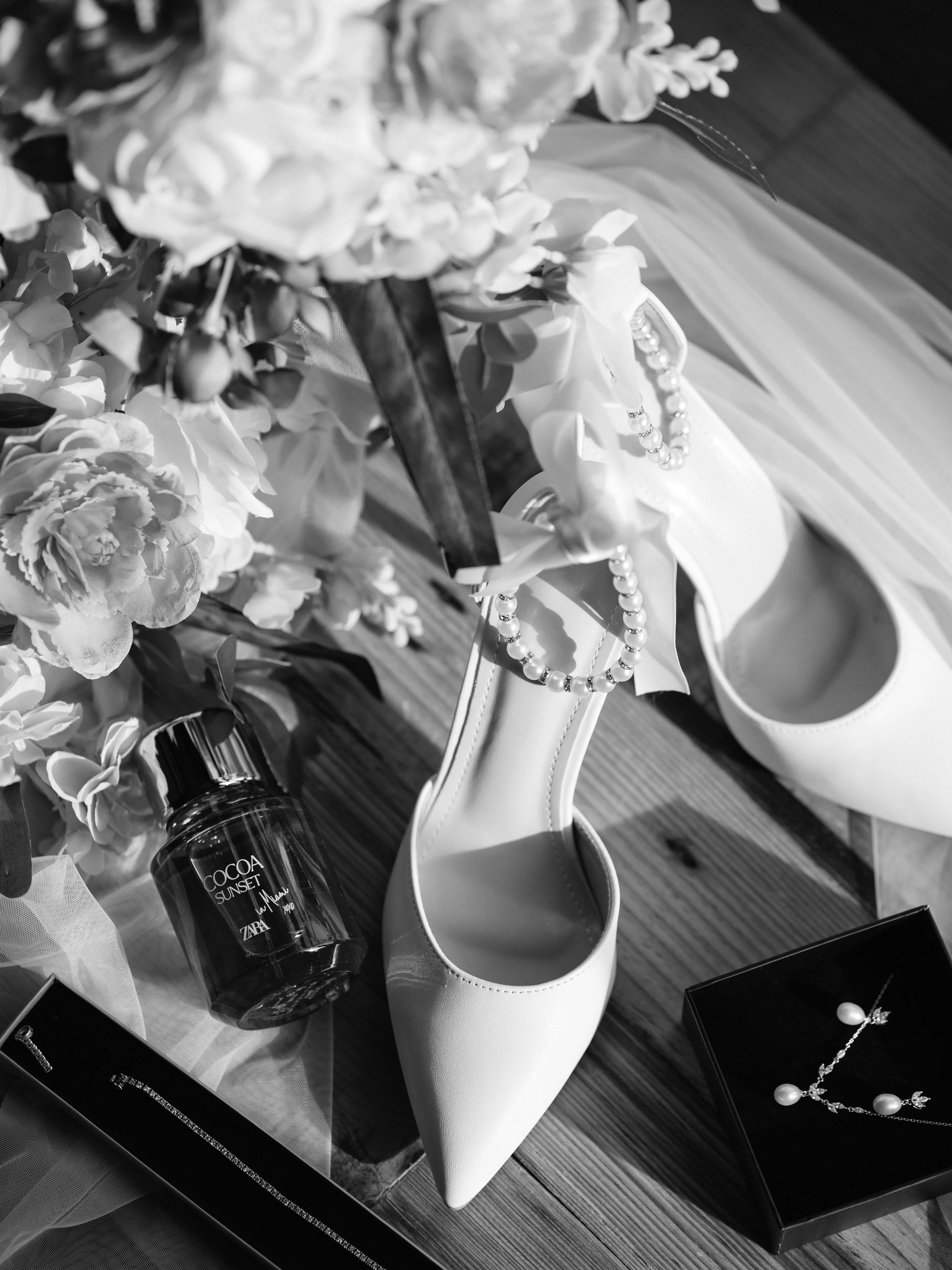 Black and white photo of various jewelry and accessories on display, including pearl necklaces, earrings, a white high-heeled shoe, a bottle labeled 'Cocoa Sunset,' and artificial flowers.