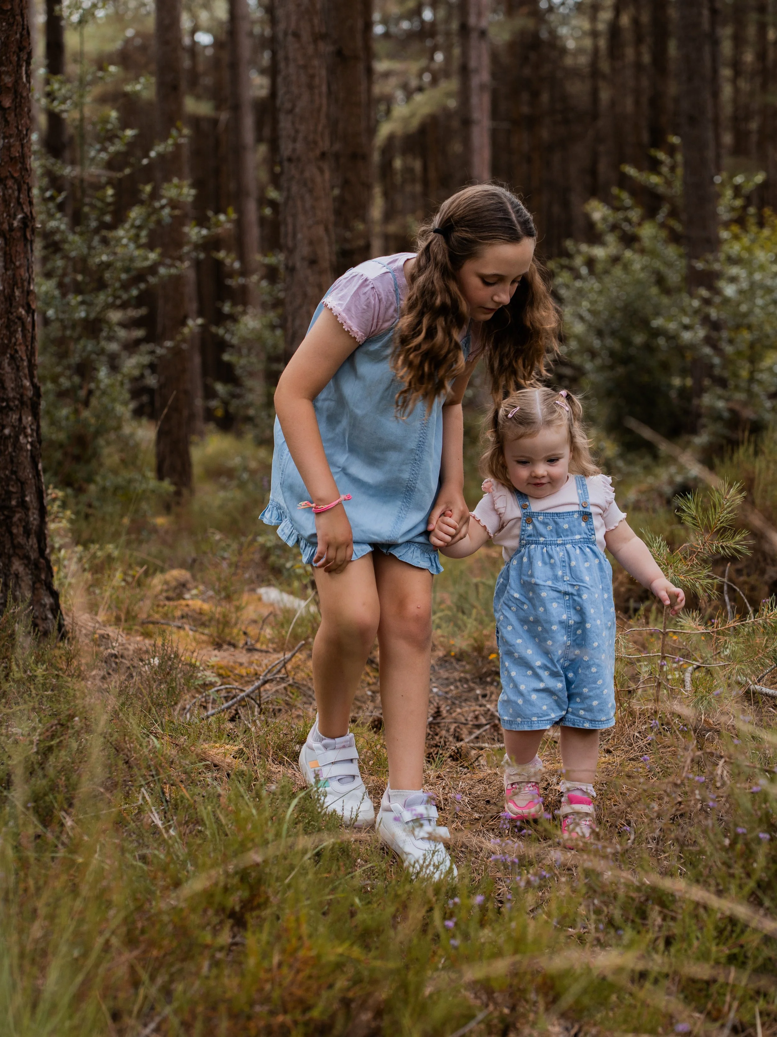 A young woman and a little girl hold hands as they walk through a forest with tall trees and green shrubs.