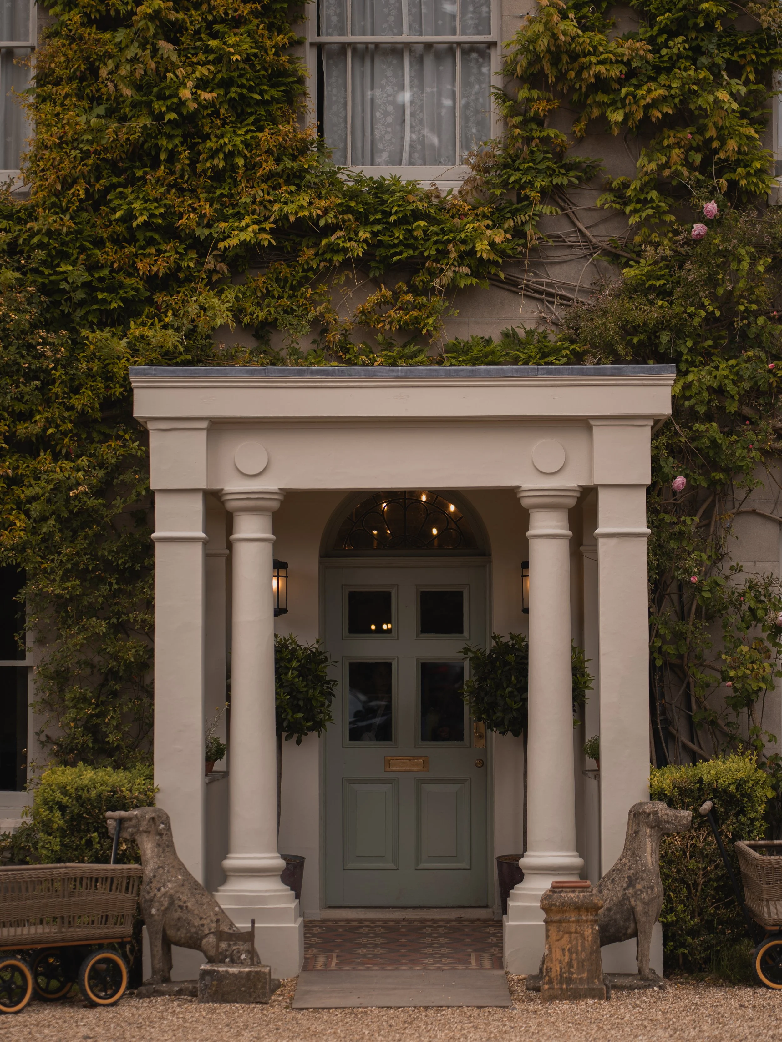 Facade of a house with a green door, white columns, surrounded by lush green vines and plants, with benches and stone sculptures in the foreground.