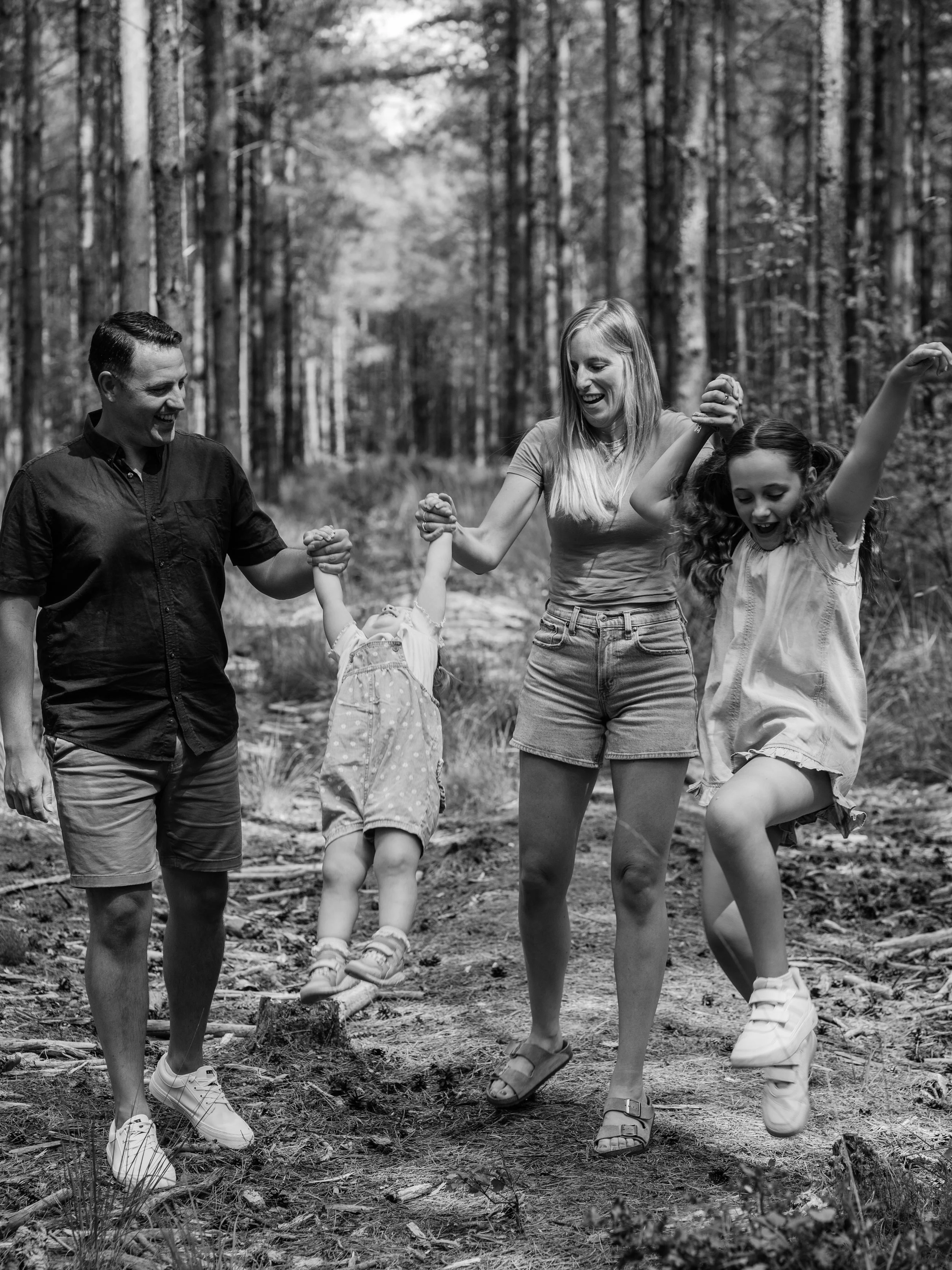 A family of four, two adults and two children, are walking and holding hands in a forest. The children are playing and the adults are holding their hands for support. The scene is outdoors with trees and natural ground.
