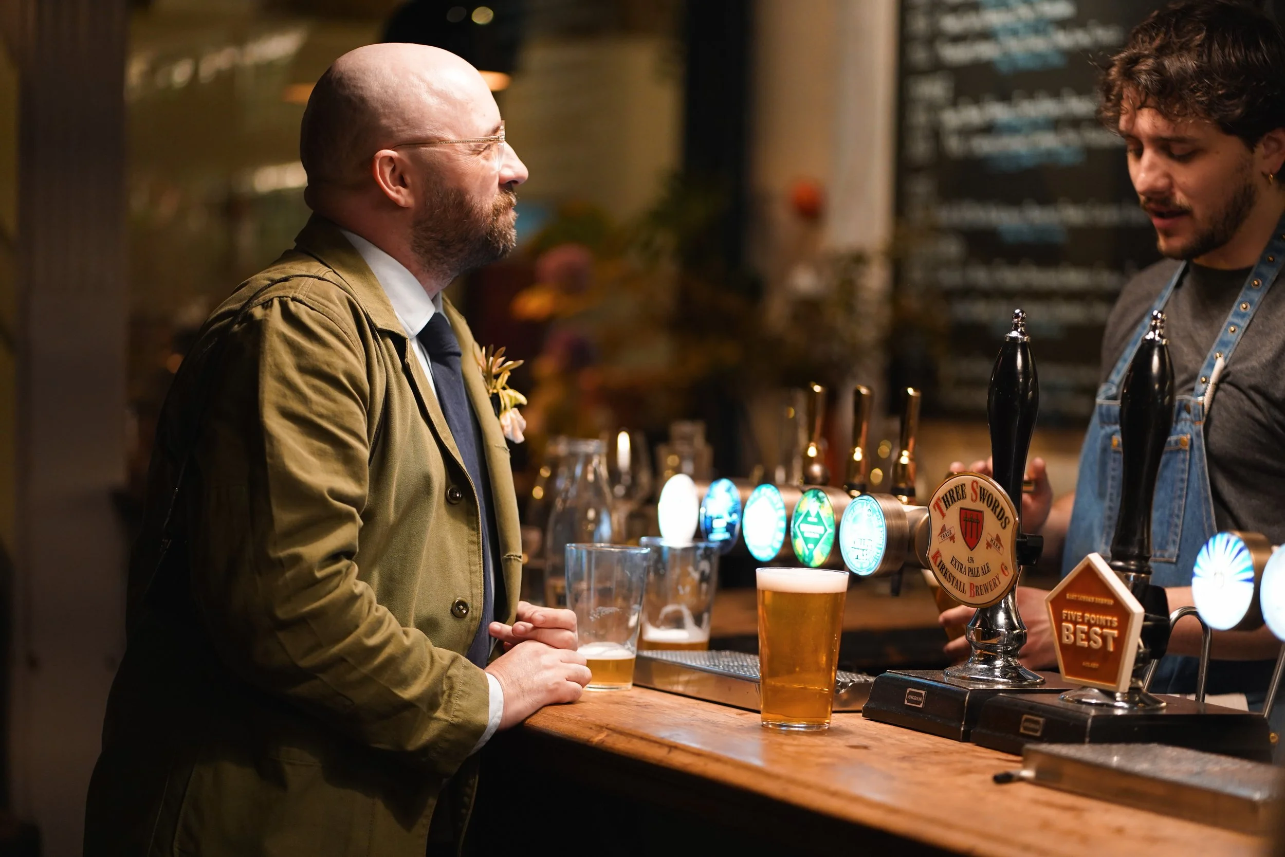A man in a green jacket and eyeglasses holding a pint of beer at a bar counter. The bartender, a young man with curly hair, is serving beer from taps with illuminated labels.