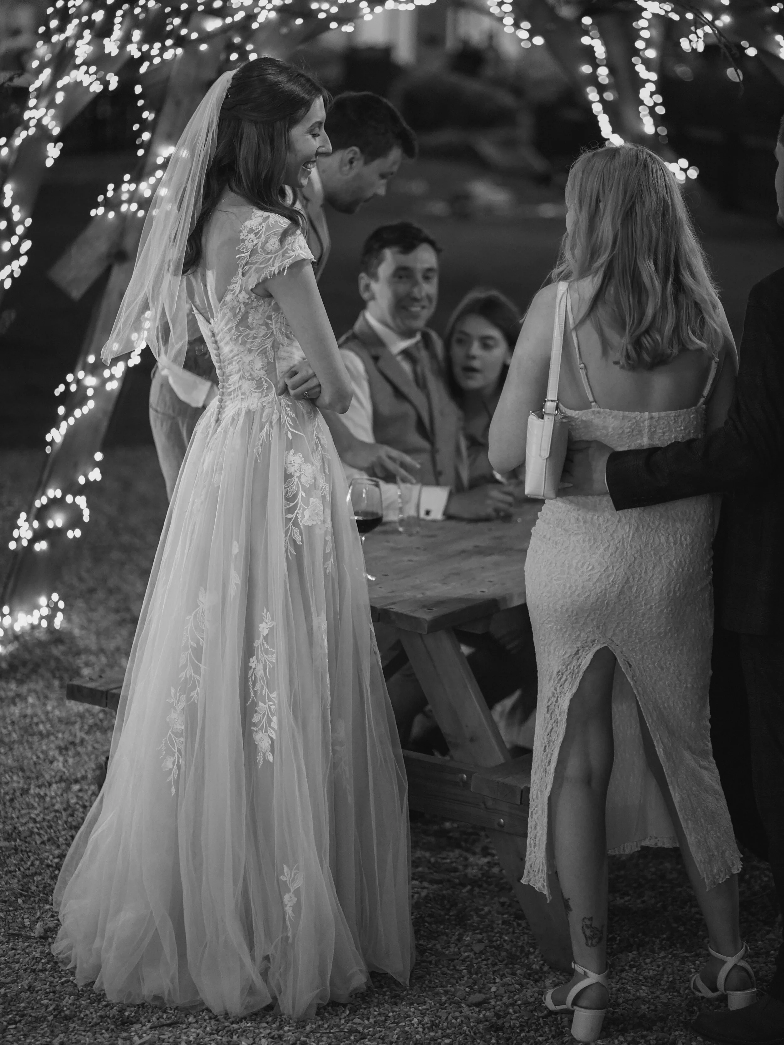 A bride in a wedding dress with lace details and a veil, smiling at guests. Guests are gathered around her under an arch decorated with string lights at an outdoor evening event.