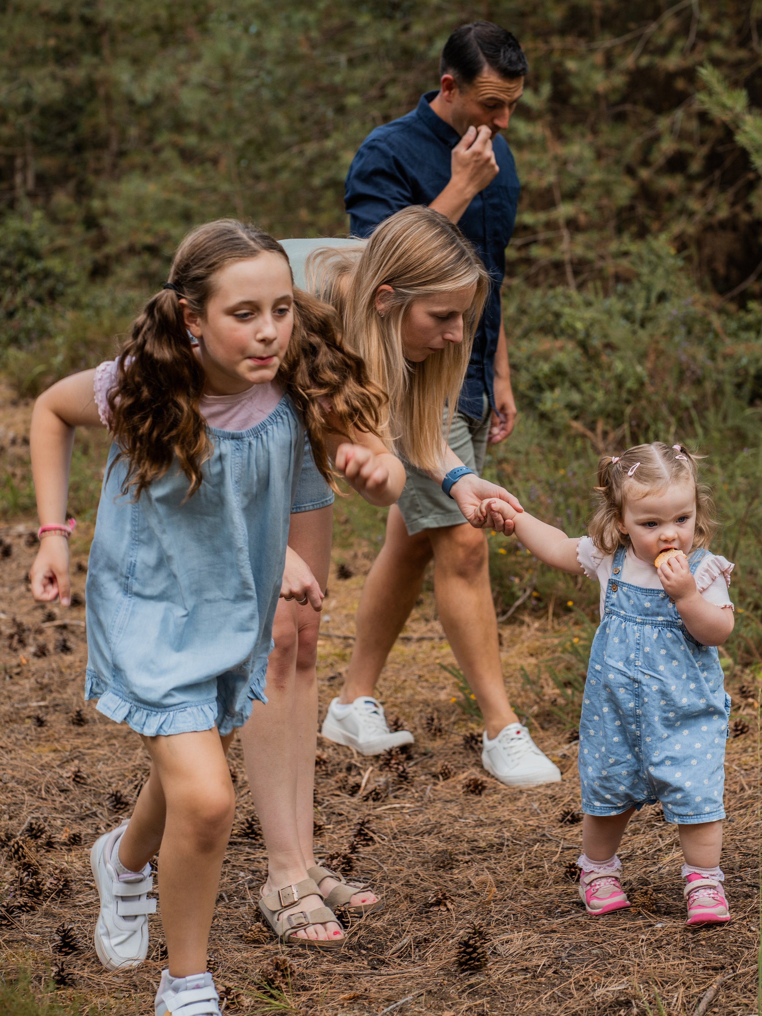A family of four children and two women hiking in a forest, holding hands and walking on a dirt trail covered with pine needles and small pine cones.