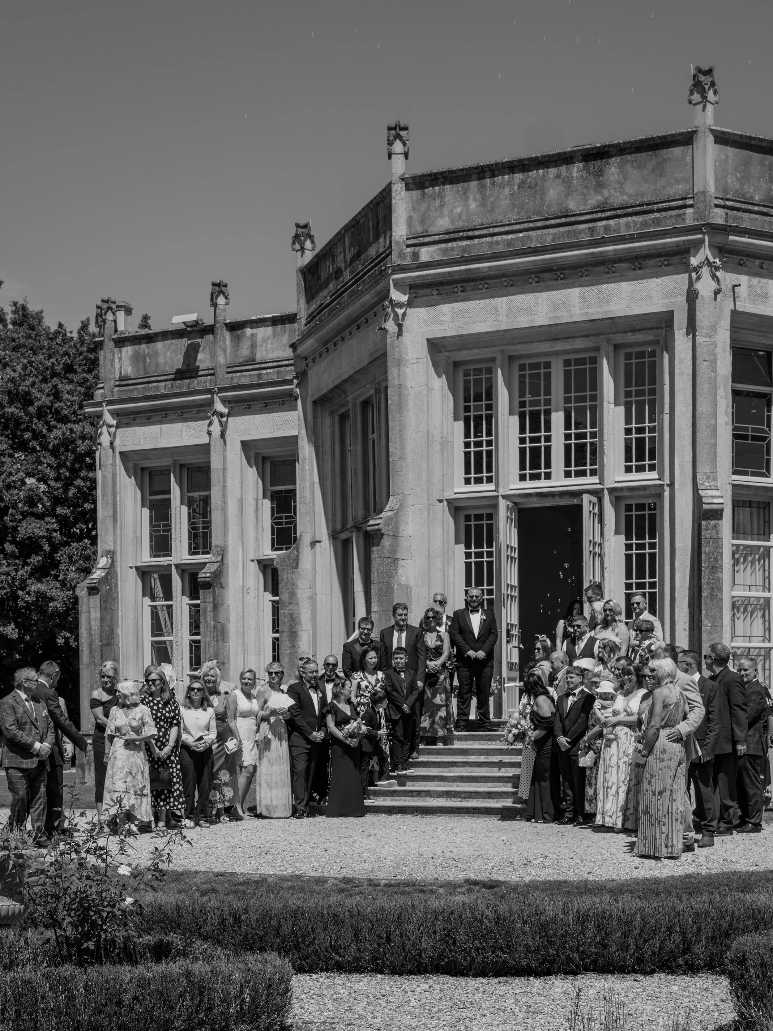 A large group of people dressed formally gathered outside a historic building with large windows and decorative elements, likely for a wedding or special event, during daytime.