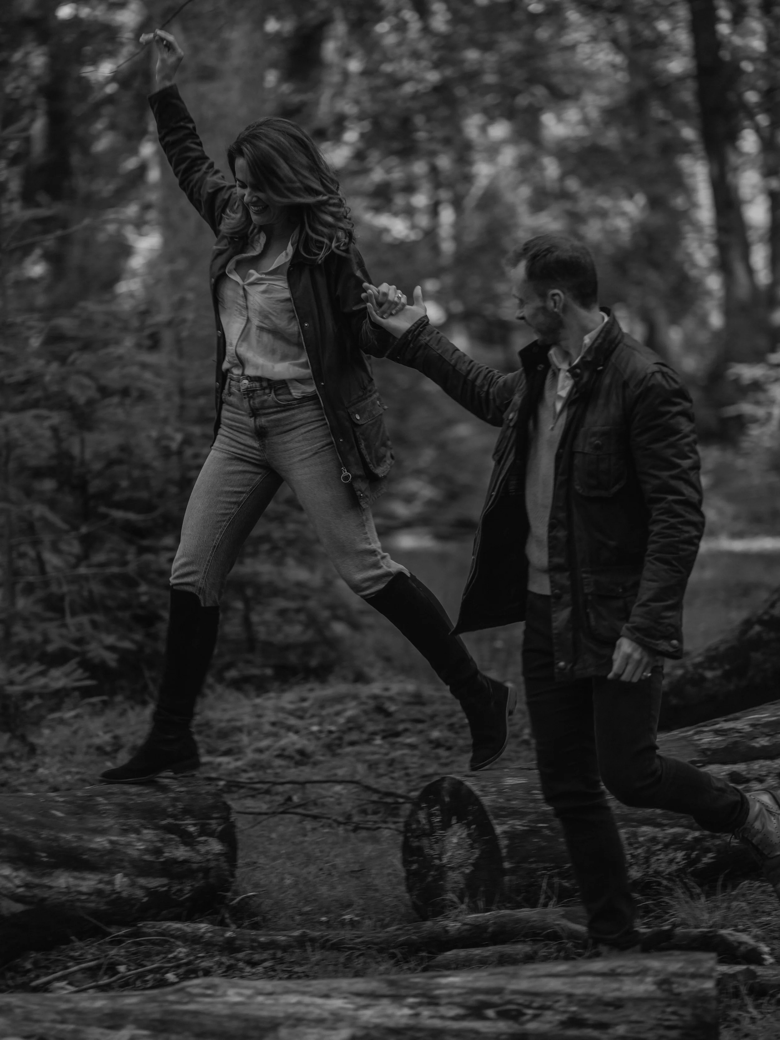 A black and white photograph of a young couple outdoors in a wooded area, holding hands and playing near a creek while smiling and enjoying each other's company.