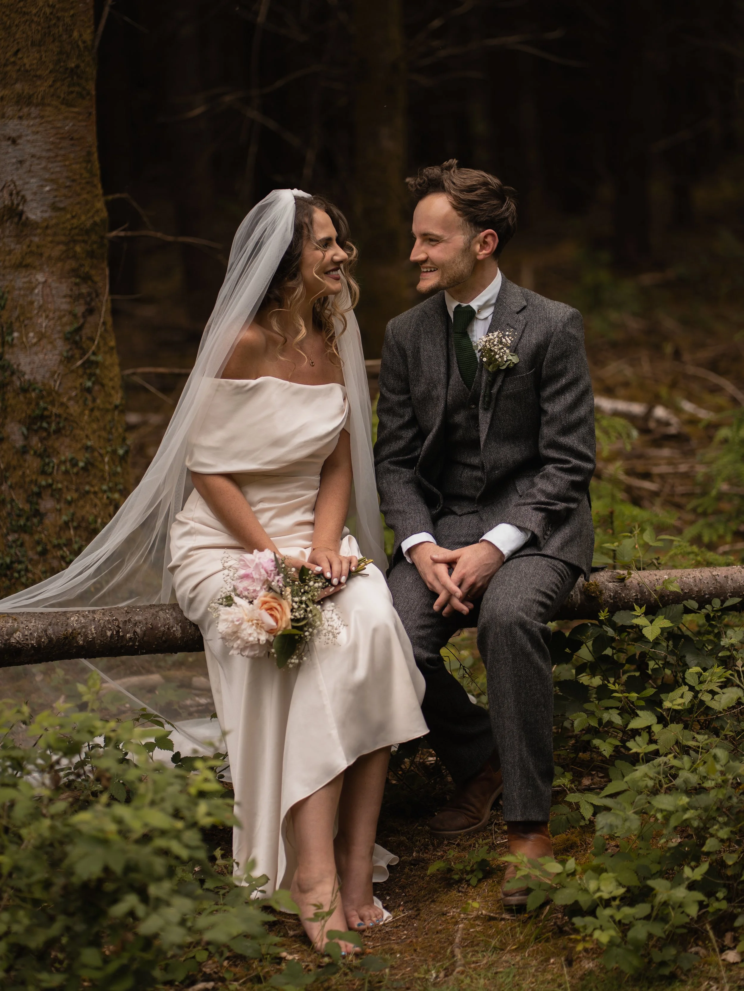 A bride with a veil and bouquet smiling at a groom in a forest setting.
